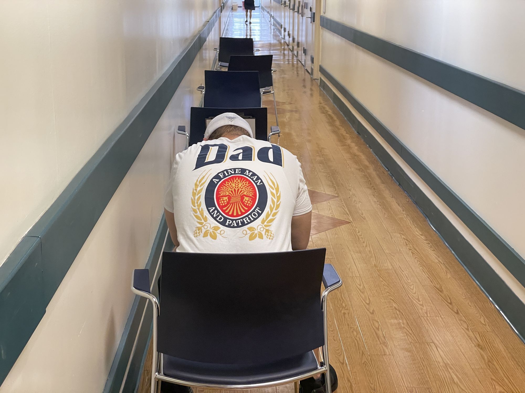Ein Mann sitzt in einem Krankenhausflur auf einem Stuhl. Er hat der Rücken zur Kamera gekehrt und seinen Kopf gesenkt. Das Wort "Dad" (Vater) ist auf seinem T-Shirt zu lesen.