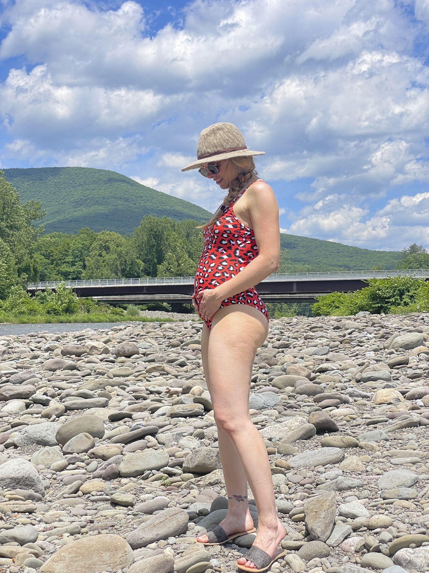 Becca steht an einem felsigen Strand, sie trägt einen Badeanzug und hält sich ihren Bauch, wie schwangere es oft tun. Es ist bereits ein kleiner Bauchansatz erkennbar.
