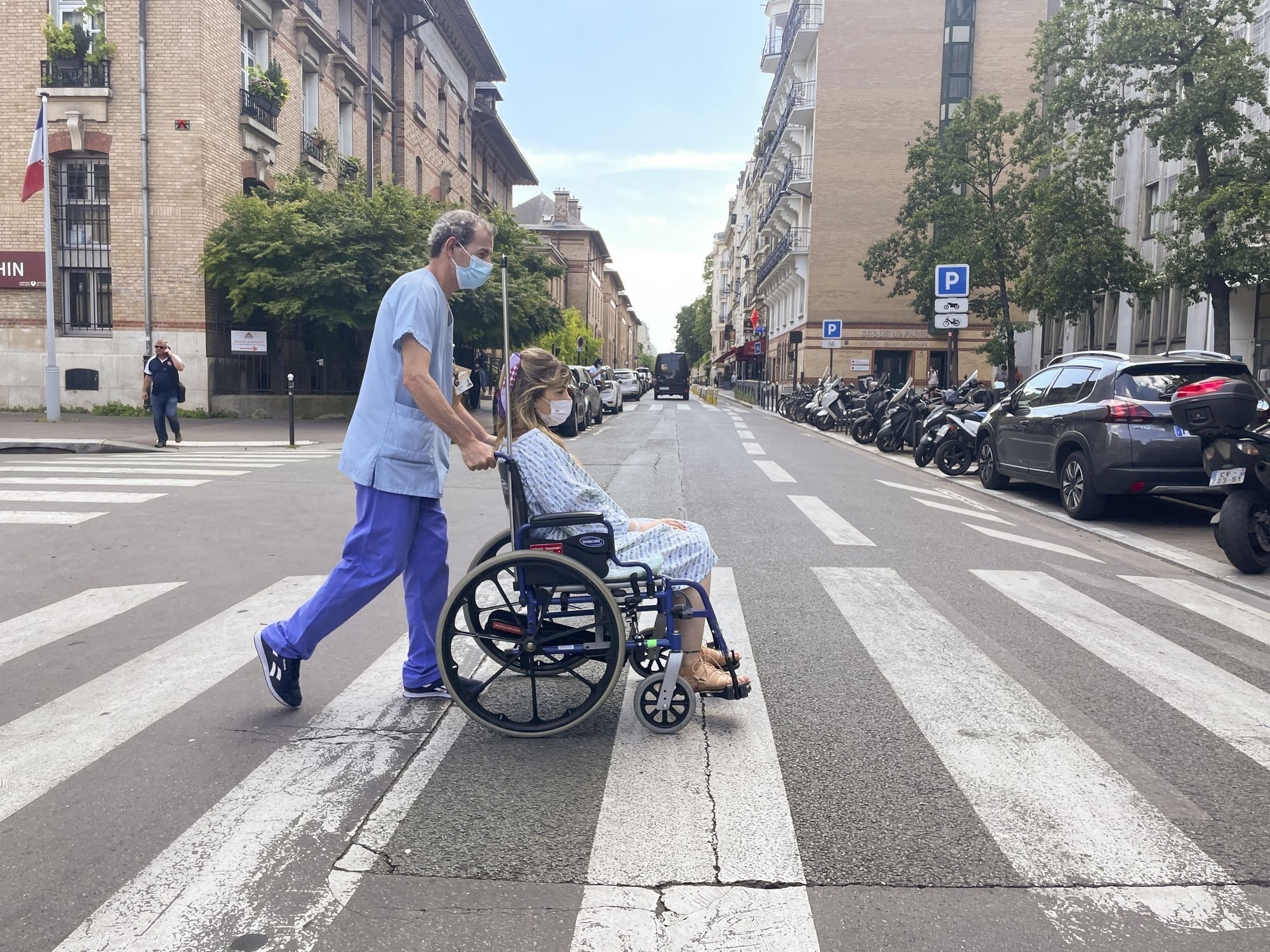Becca wird von einem Krankenpfleger in einem Rollstuhl über den Zebrastreifen geschoben. Sie trägt einen Krankenhauskittel.
