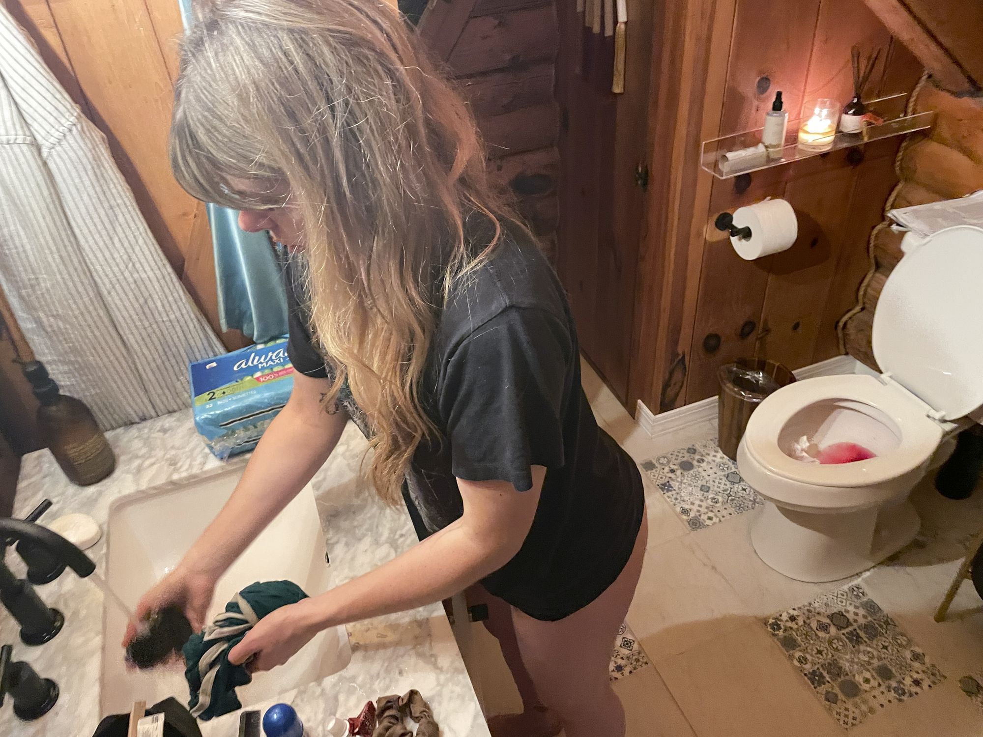 Becca steht im Badezimmer und wäscht ein Kleidungsstück im Waschbecken aus. Im Hintergrund ist die Toilette zu sehen, in ihr befindet sich Blut.