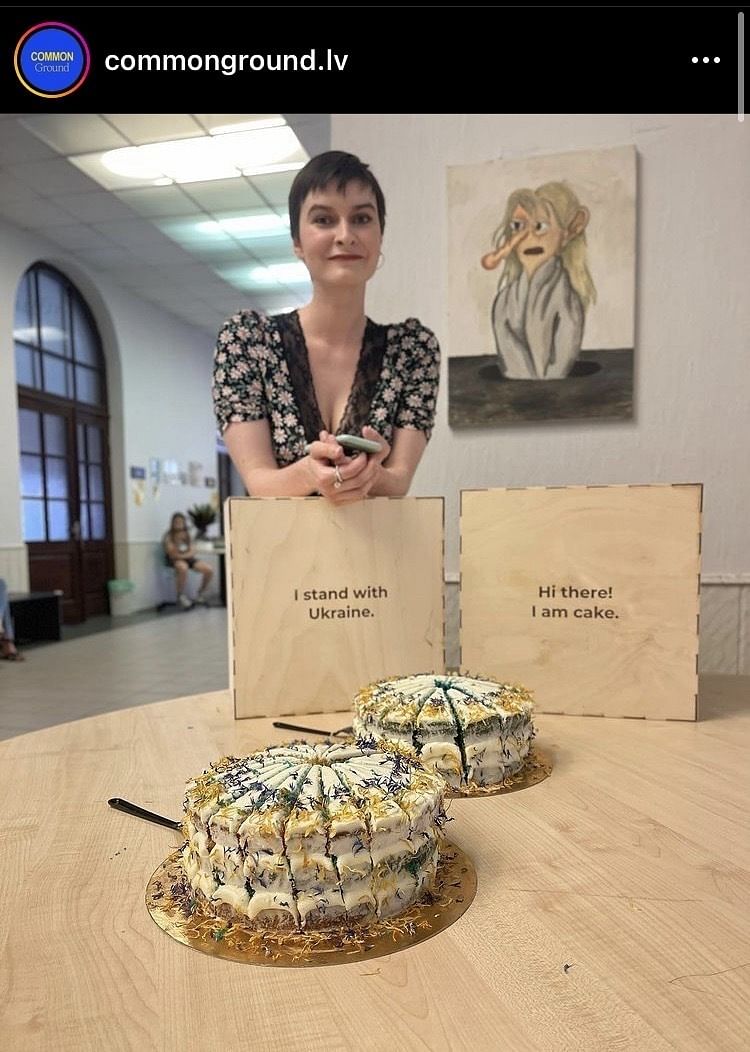 Eine Frau mit kurzen Haaren Steht lehnt sich auf eine Kuchenbox, auf der steht "I stand with Ukraine". Davor stehen zwei Torten in den Nationalfarben der Ukraine.