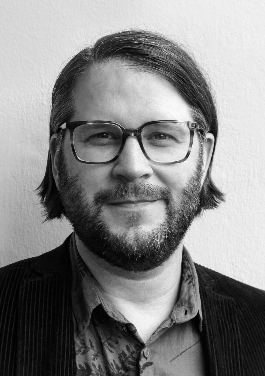 Auf dem schwarz-weißen Foto ist ein Mann mittleren Alters zu sehen. Er hat mittellanges Haar und trägt eine Brille und einen gestutzten Vollbart.