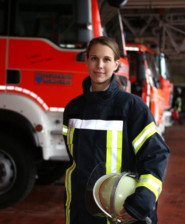 Eine Frau blickt in die Kamera, sie trägt eine Feuerwehr-Uniform. Hinter ihr sind Feuerwehrautos zu sehen. Ihren Helm trägt sie unter dem Arm.