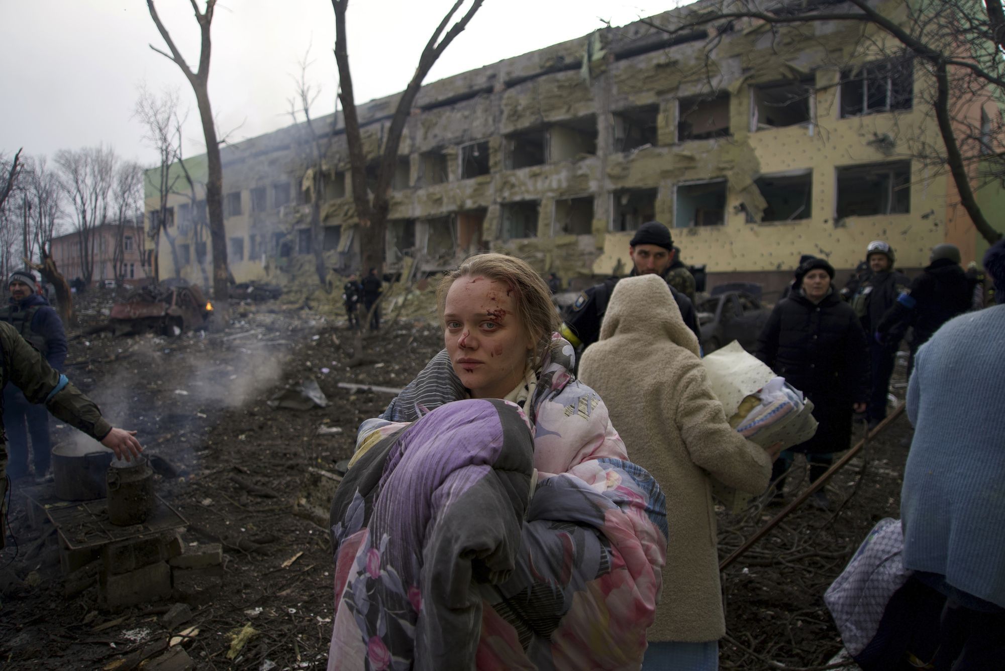 Eine Frau steht vor der zerbombten Geburtsstation, sie ist in eine Decke gehüllt und hat eine Wunde am Kopf. Sie blickt in die Kamera. Hinter ihr laufen Menschen entlang.