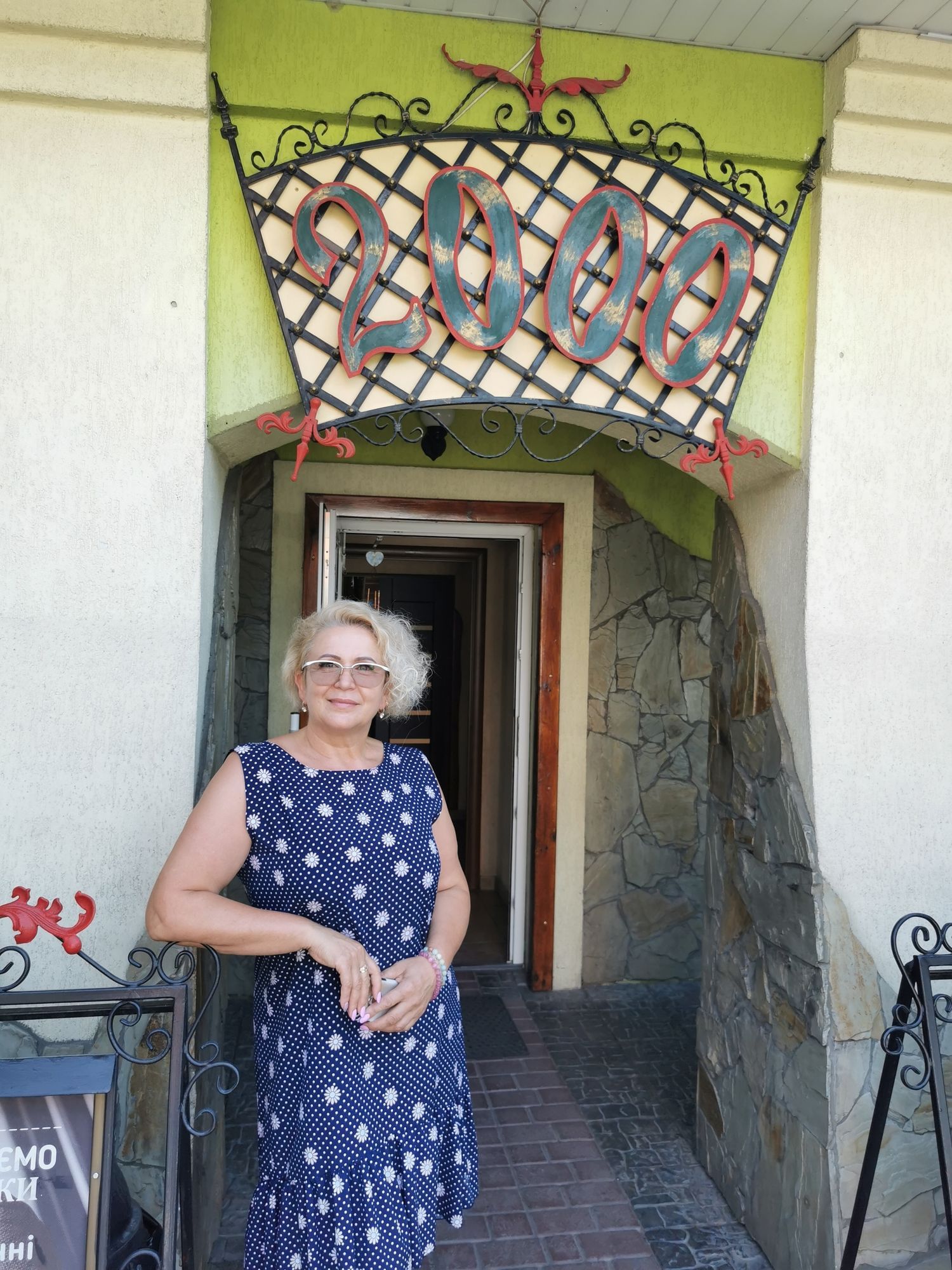 Eine blonde Steht vor einem Eingang, über dem Eingang steht "2000". Sie trägt ein blaues Kleid mit weißen Punkten.