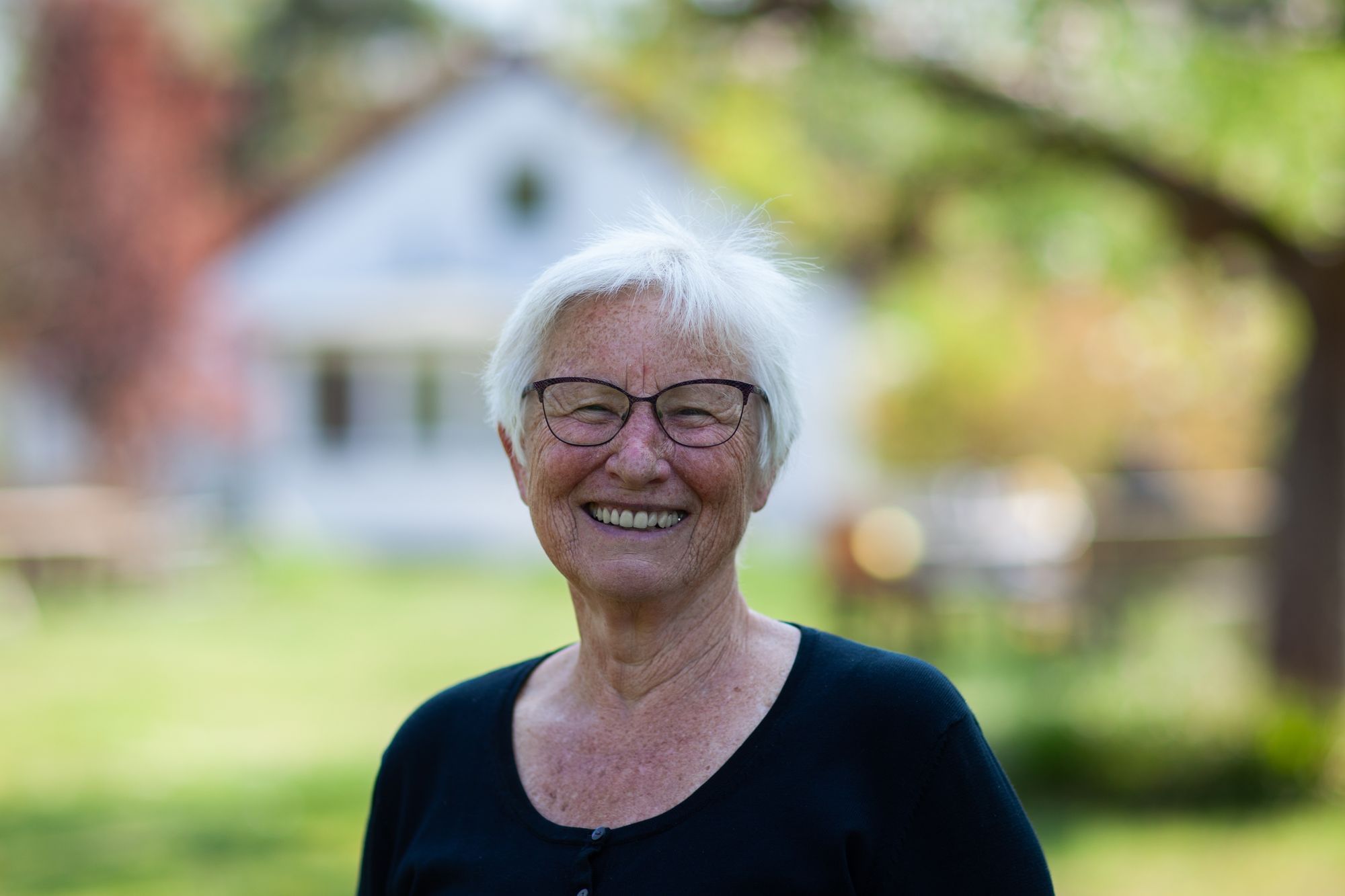 Wir sehen eine lächelnde Frau mit grauen Haaren, die vor ihrem Haus und einem Baum steht.