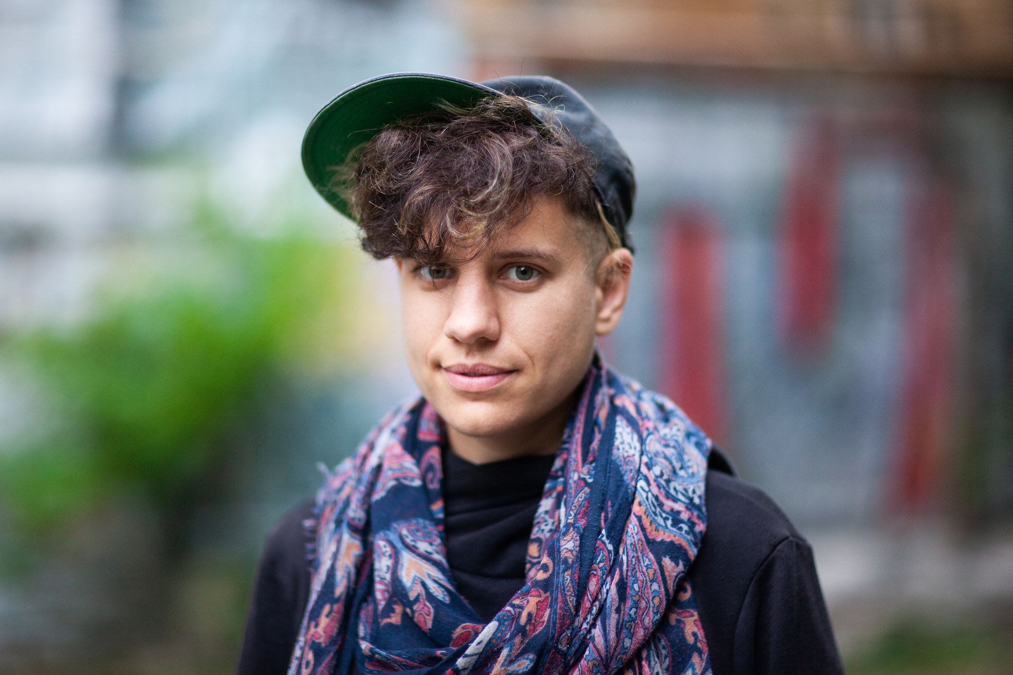 Wir sehen eine junge Person mit Locken und einer Kappe, die in die Kamera schaut.