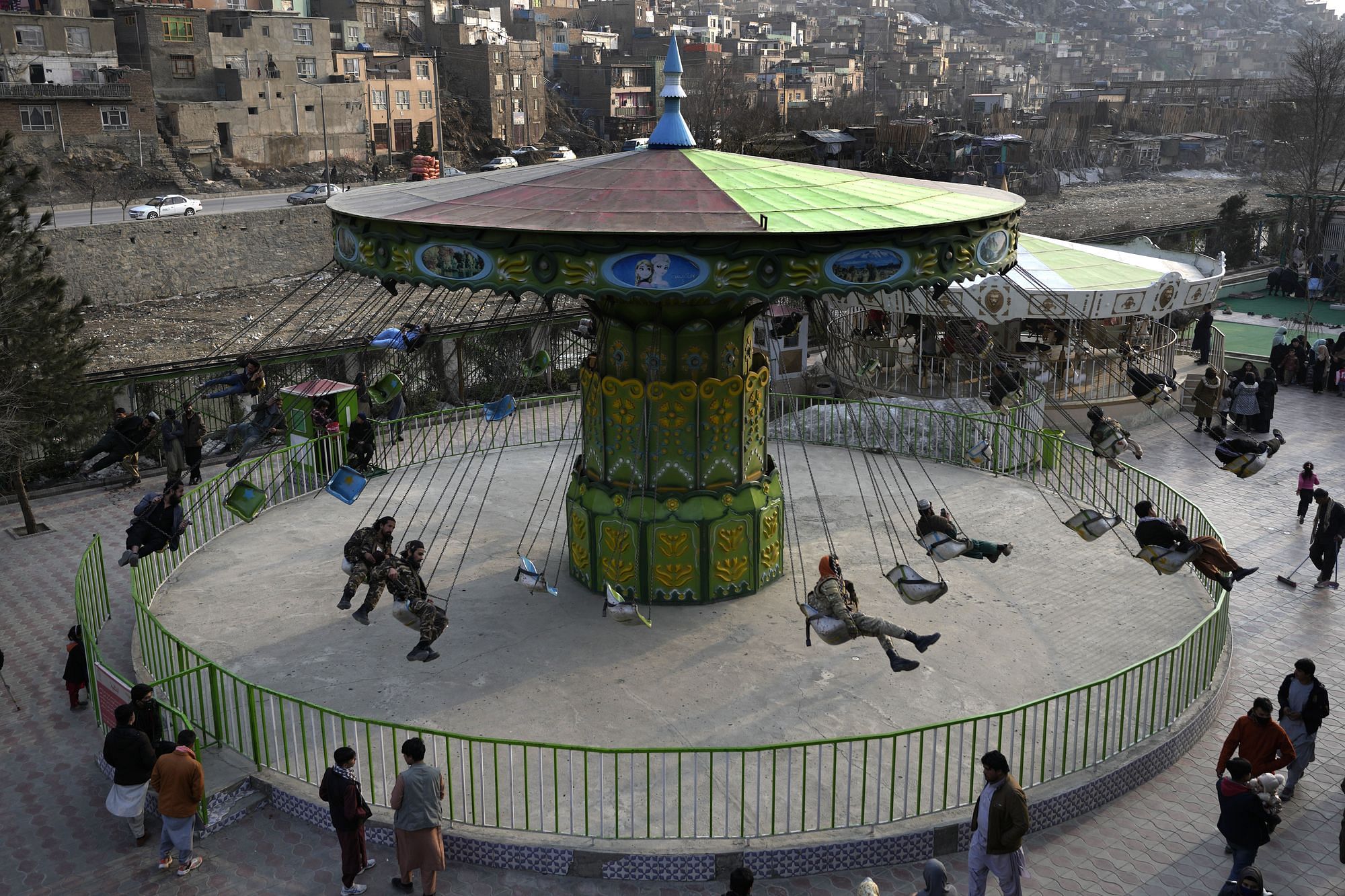 Aufnahme eines Kettenkarussells von leicht oben. Männer belegen die Sitze und schwingen daran. Einige Leute stehen davor und sehen zu. Im Hintergrund ist Kabul zu sehen.