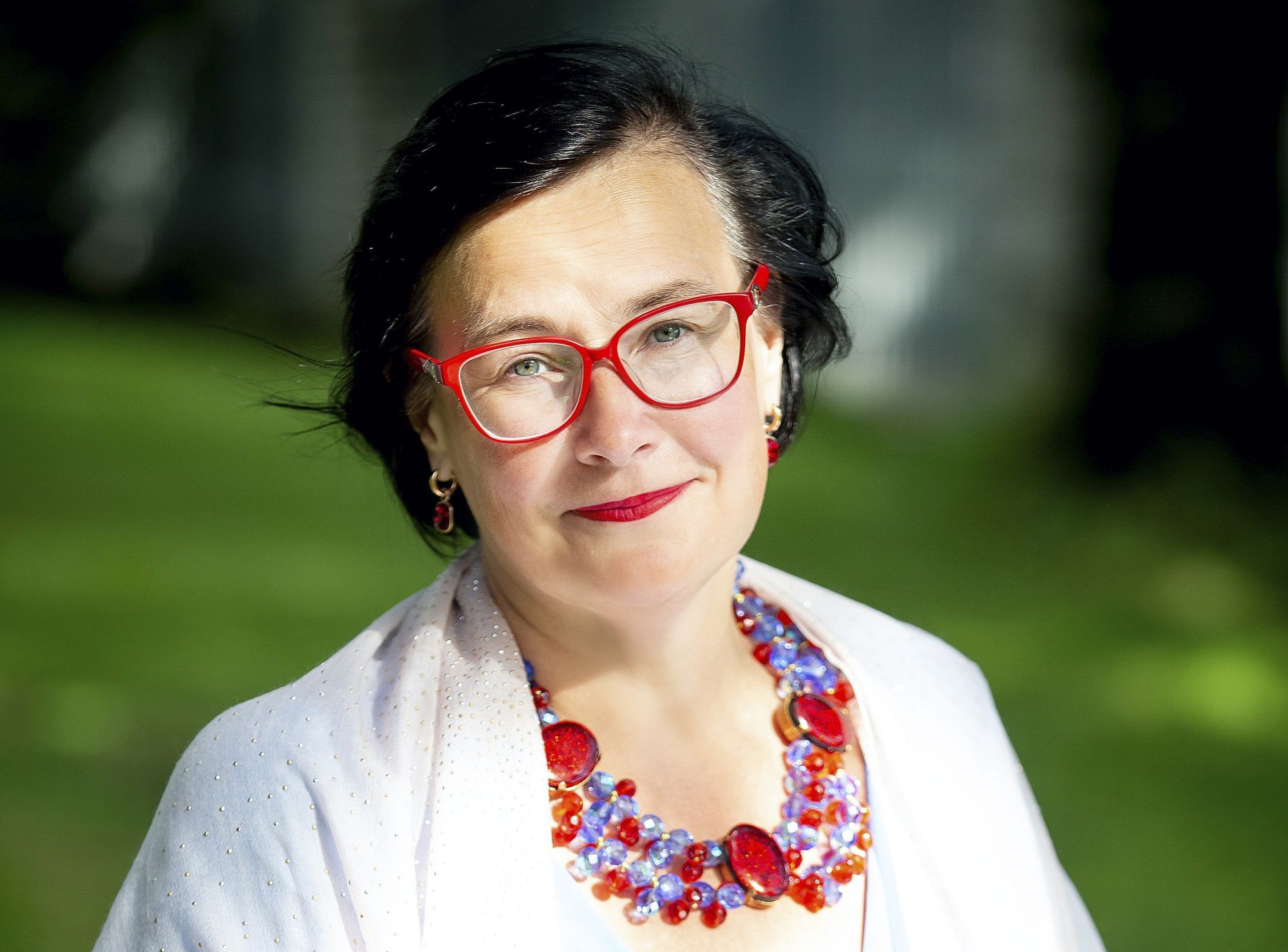 Eine Frau mittleren Alters, sie trägt eine weiße Bluse, eine rote Brille und eine dazu passende Kette aus rotem und blauem Glas.