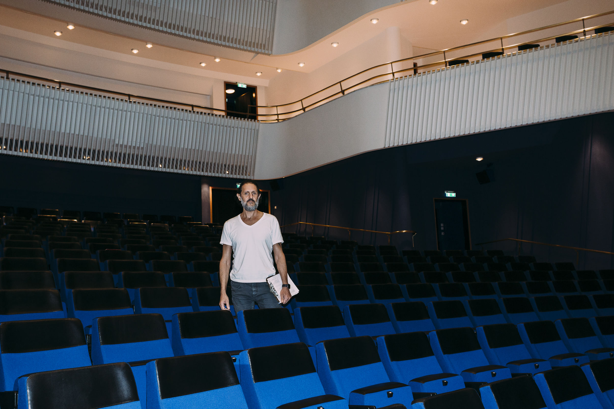 Saad steht zwischen den Sitzen des Theaters. Er hält sein Klemmbrett in der Hand.