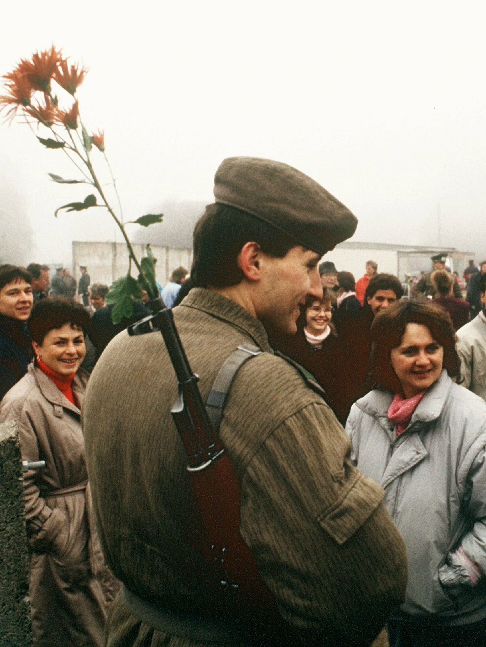 Ein DDR-Soldat steht umringt von Zivilisten. Er trägt ein Gewehr geschultert, eine rote Blume steckt im Lauf.