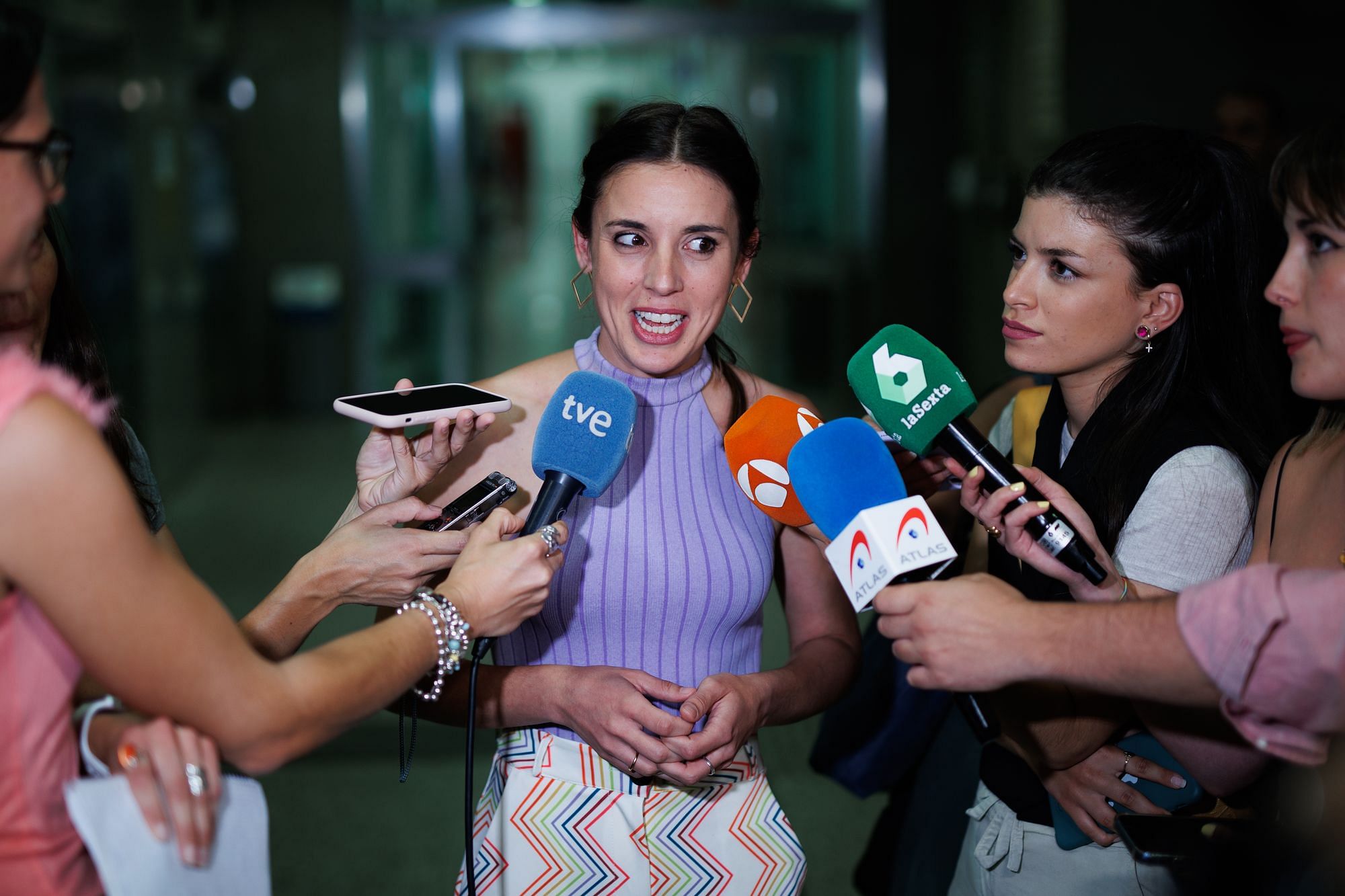Man sieht die Politikerin Irene Montero umringt von Journalistinnen, die ihr Mikros vors Gesicht halten. Mentero trägt ein schulterfreies, helllila Top und eine farbige Hose, sie wirkt damit sehr leger und gar nicht wie eine Politikerin.