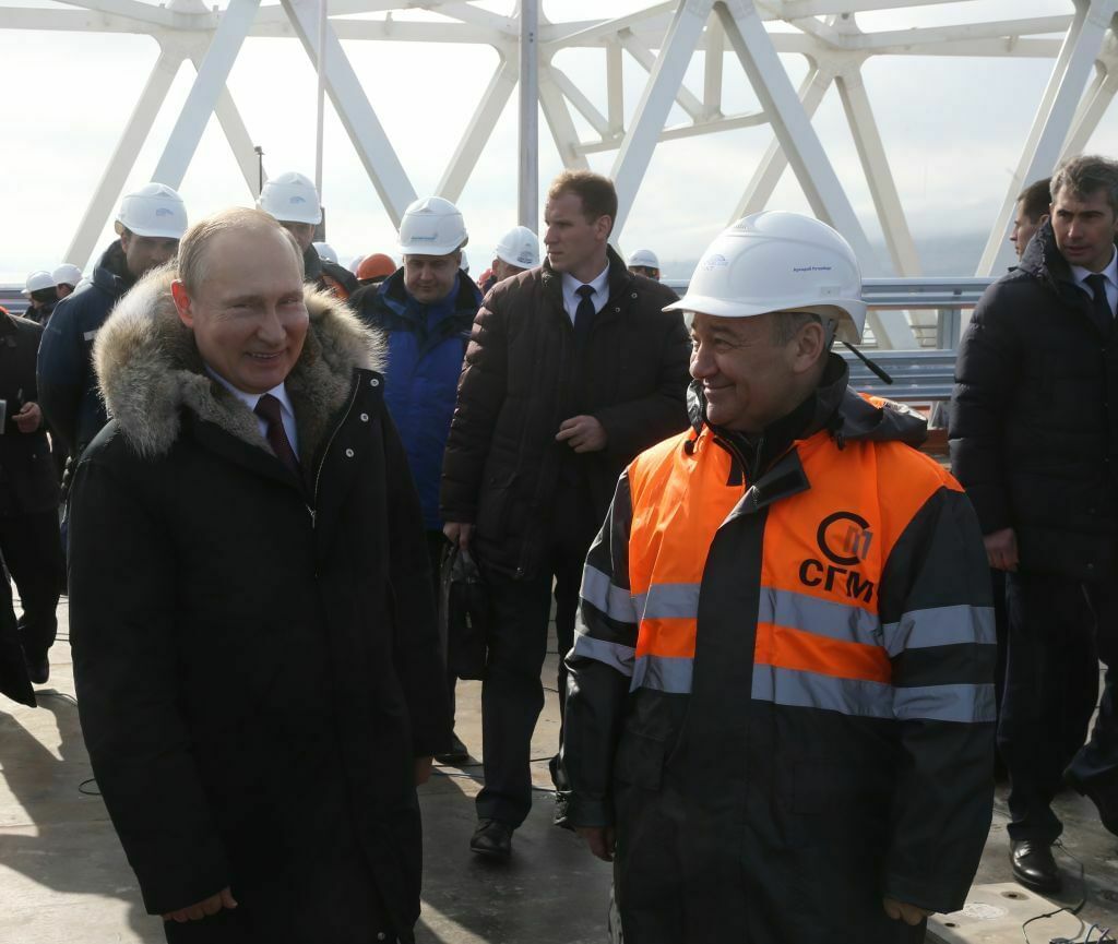 Der russische Präsident Vladimir Putin trägt eine dicke Winterjacke mit Pelz. Neben ihm steht ein Mann mit Baustellenhelm und orangefarbener Weste. Sie grinsen sich schelmisch an.