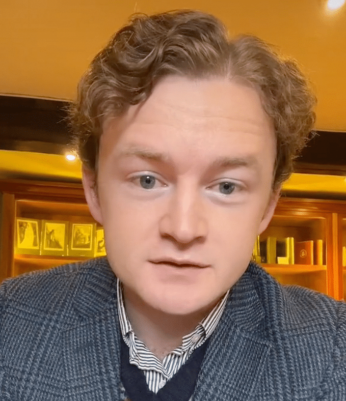 Man sieht einen jungen Mann mit hoher Stirn und gewellten blonden Haaren in Jacket und Hemd, wie er in eine Kamera guckt, sehr nah. Das ist Tom Wayling.