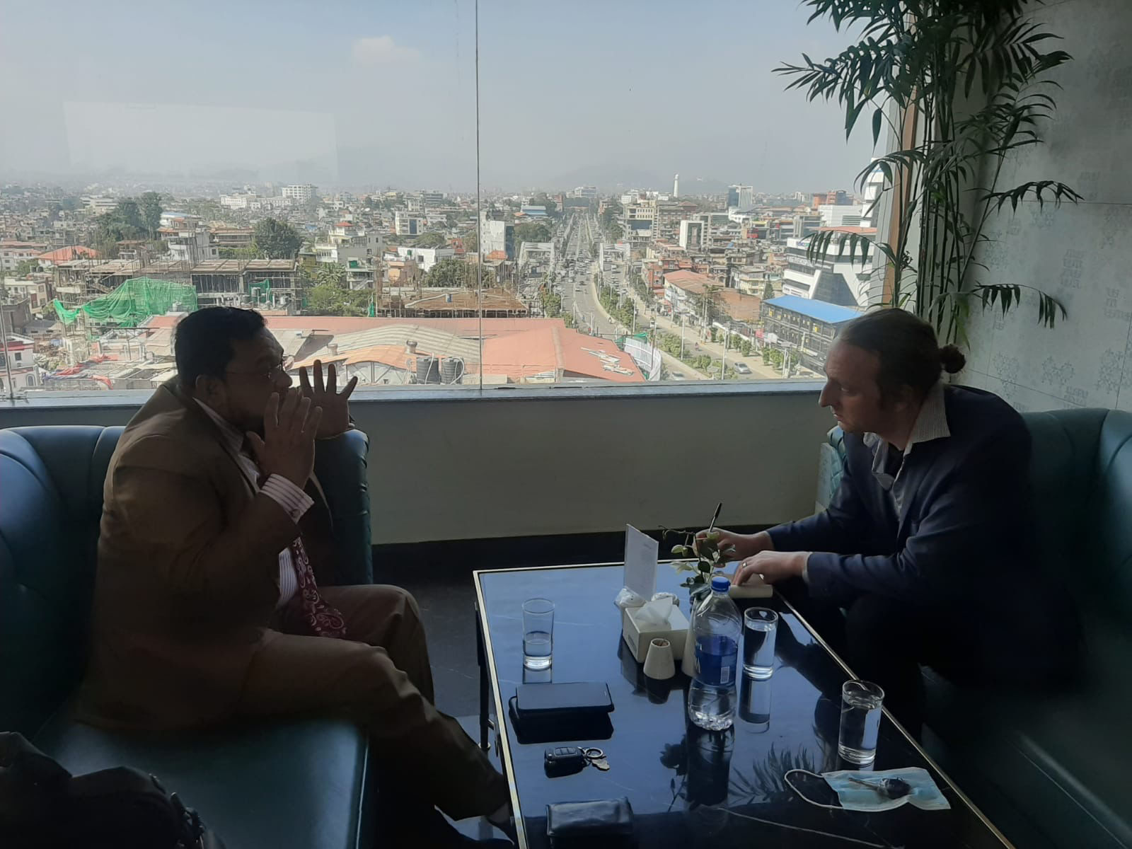 Zwei Männer sitzen sich gegenüber und unterhalten sich. Im Hintergrund ist eine Stadt durch ein Fenster zu erkennen.