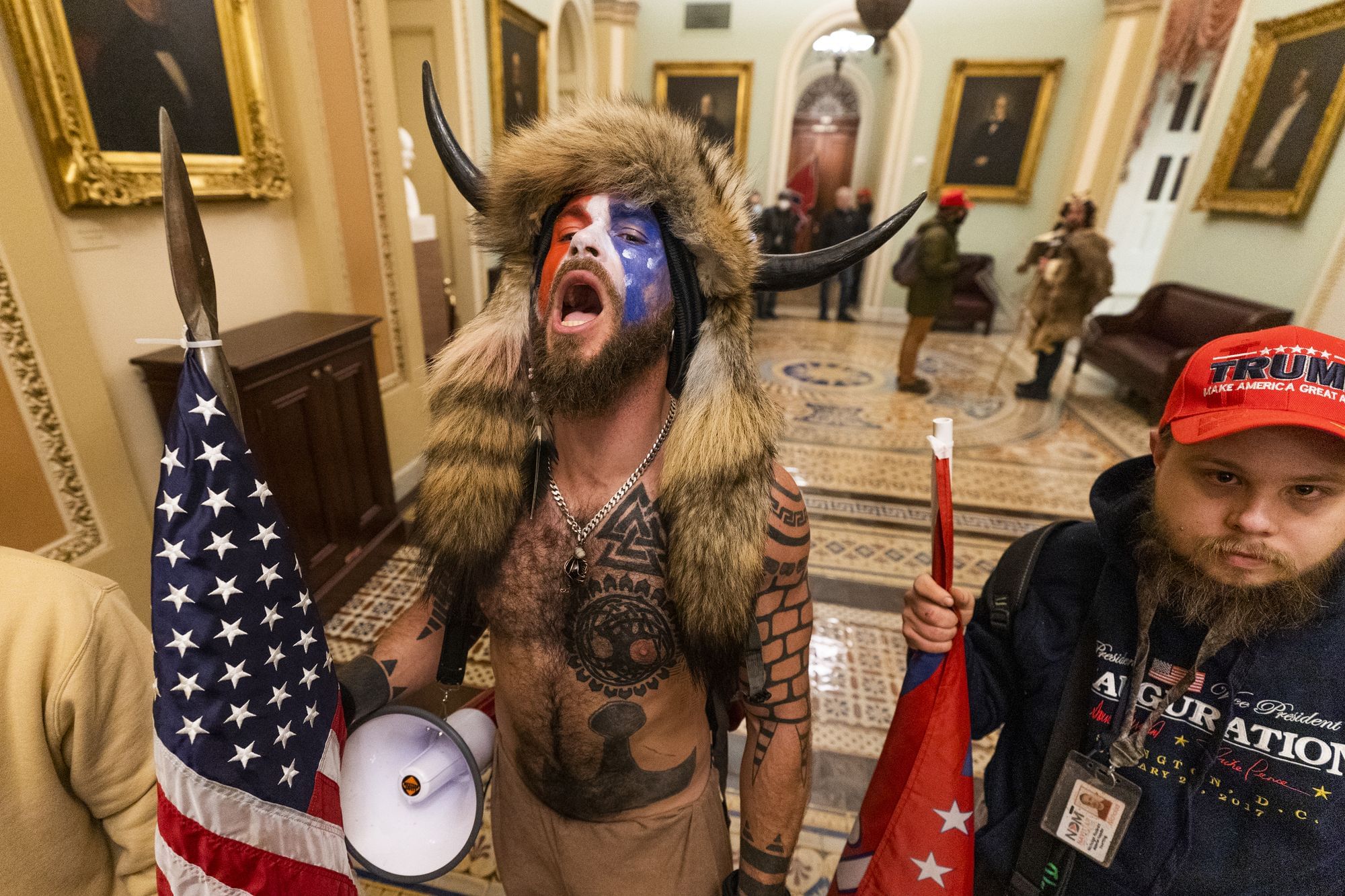 Ein als Schamane verkleideter Mann steht schreiend im Kapitol. Er hält einen Speer, an dem eine USA-Flagge befestigt ist.