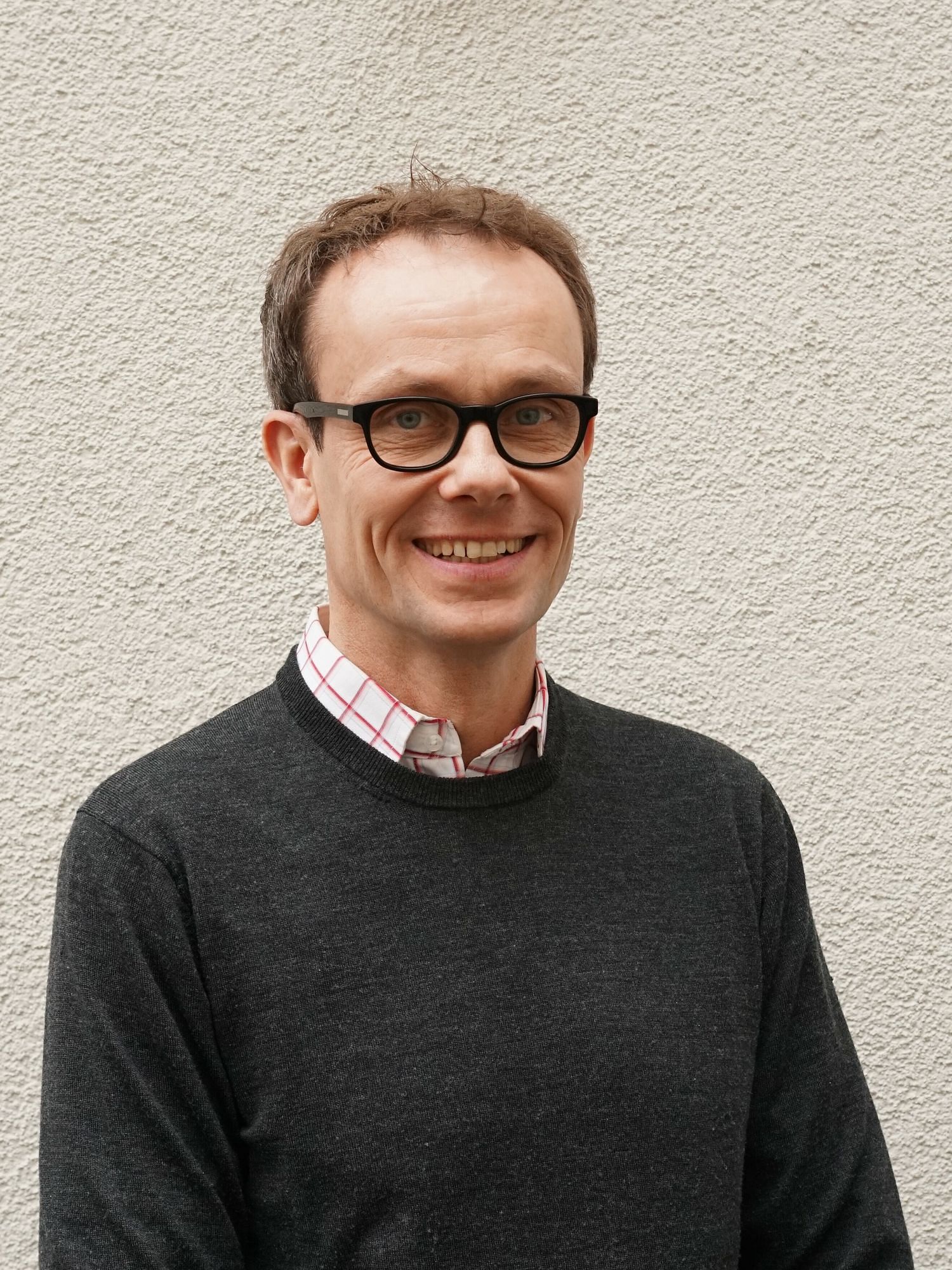 Wir sehen Tom Bschor, ein Mann mit Brille und kurzem Haar, der vor einer Hauswand in die Kamera lächelt. 