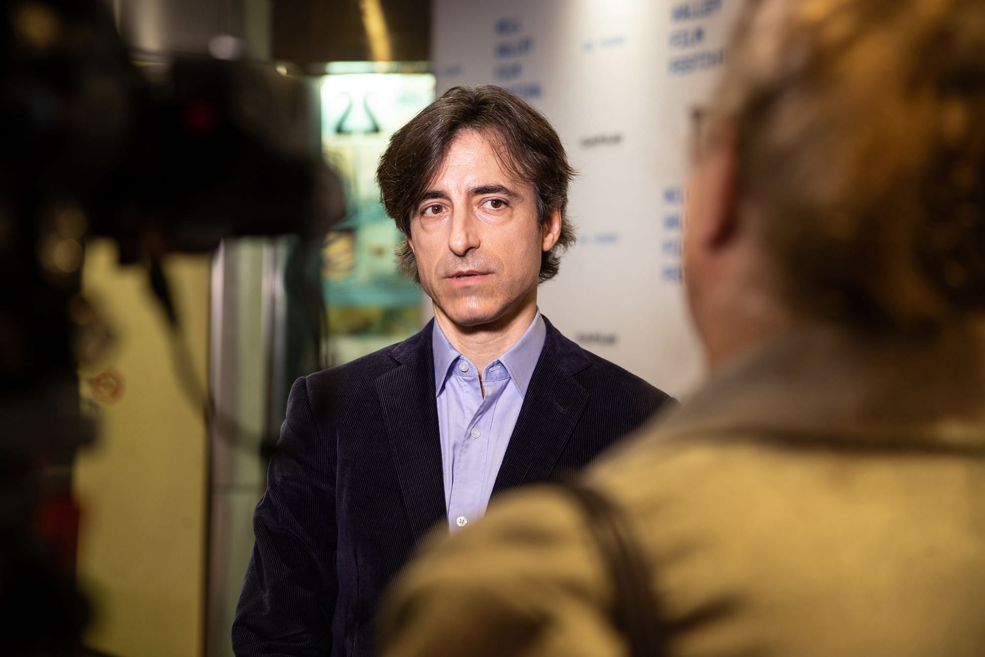 Man sieht Noah Baum, wie er auf einer Pressekonferenz interviewt wird. Schwarzes kurzes Haar, lavendelfarbenes Hemd, schwarzes Sakko