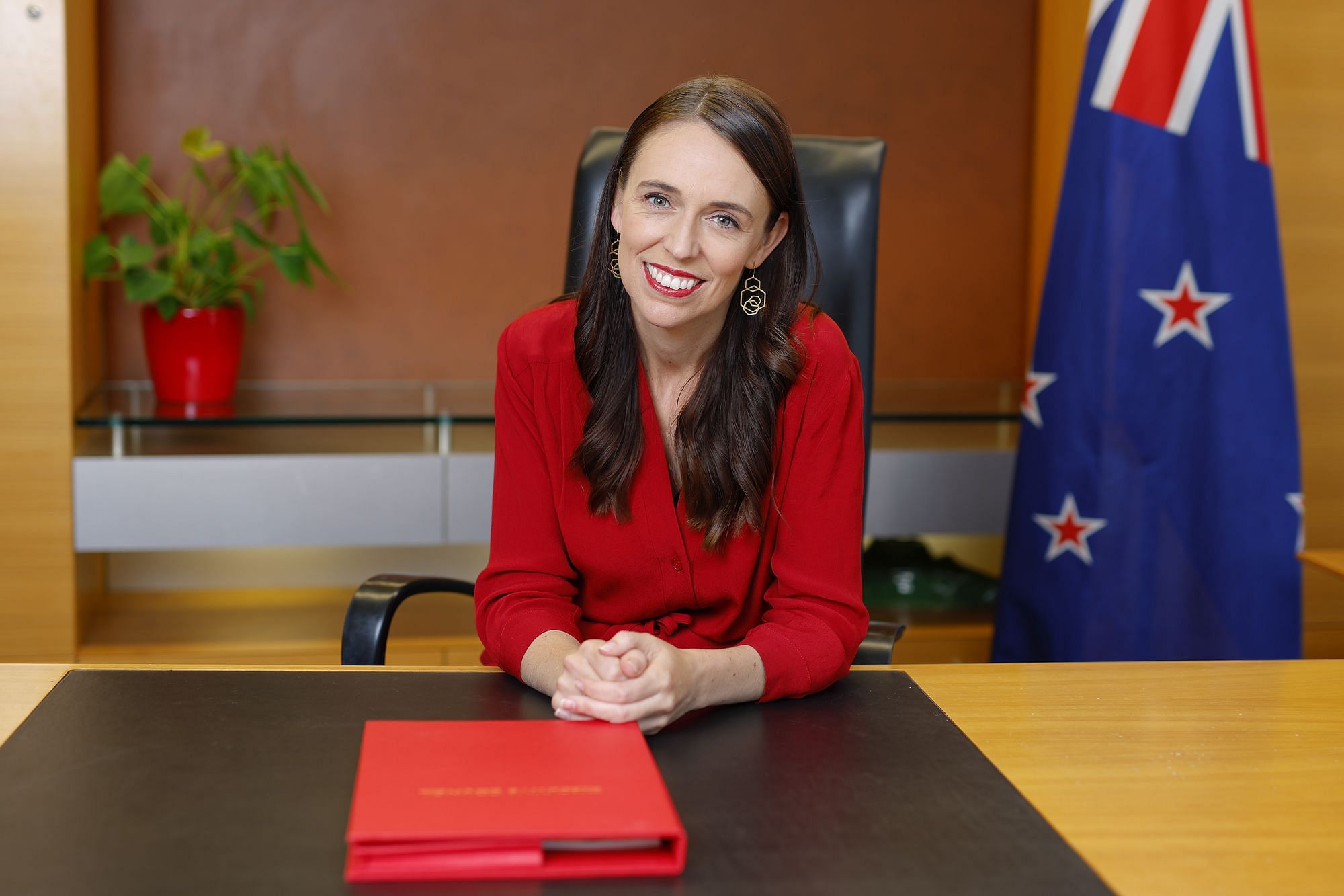 Jacinda Ardern sitzt lächelnd an einem Schreibtisch. Sie trägt eine rote Bluse, die Lippen sind rot geschminkt. Im Hintergrund steht rechts die neuseeländische Flagge.