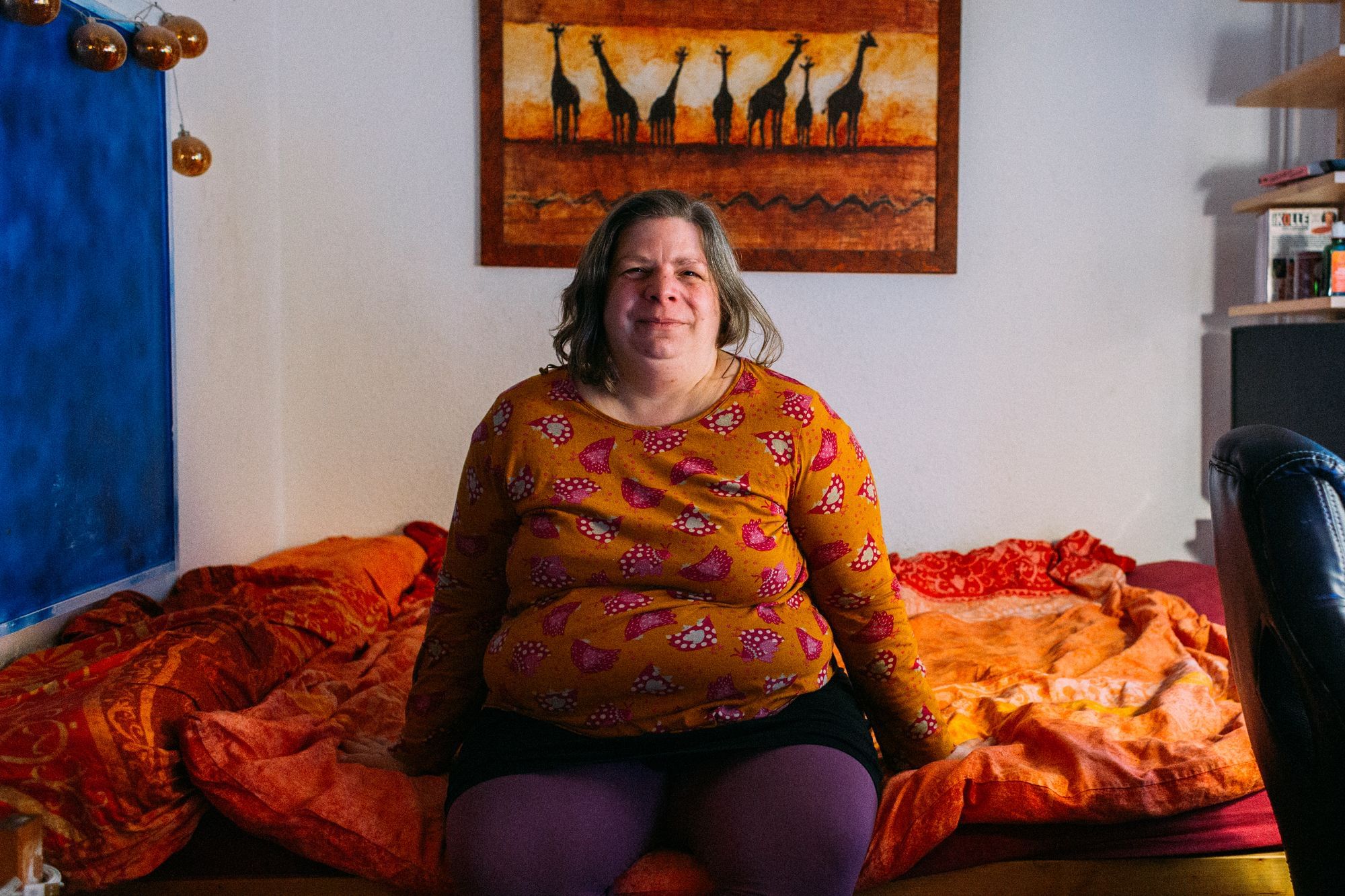 Zu sehen ist Susa, die auf einem Bett sitzt, alles ist orange: Susas T-Shirt, die Bettwäsche, das Bild von Giraffen über dem Bett.
