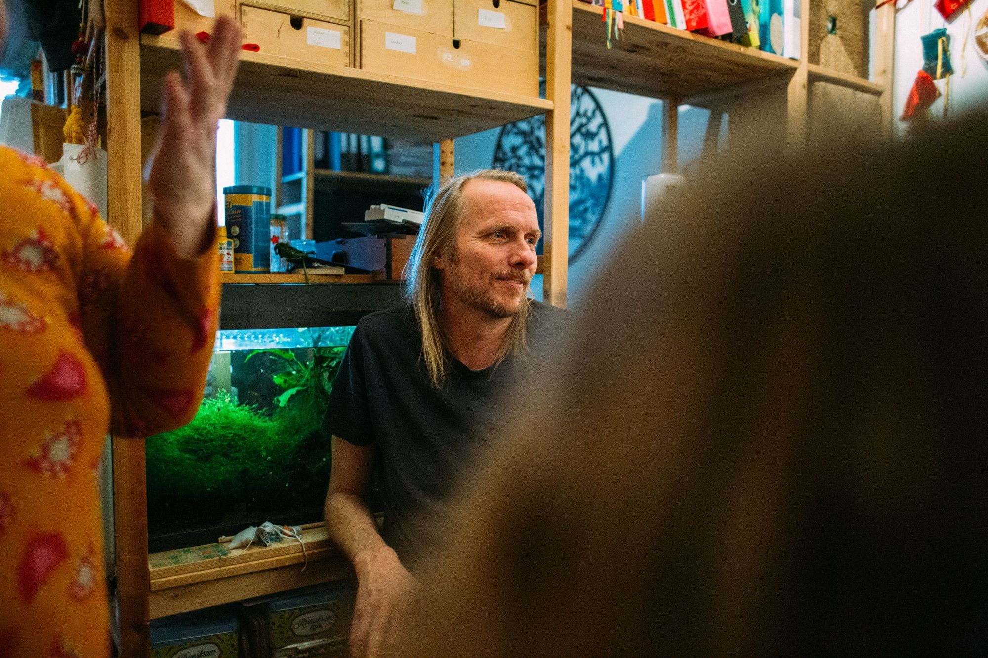 Vor einem kleinen Aquarium sitzt Arne, ein schlanker, blonder Mann. Er schaut nach rechts. Zu sehen ist auch Susas Arm und ein Kopf im Vordergrund.