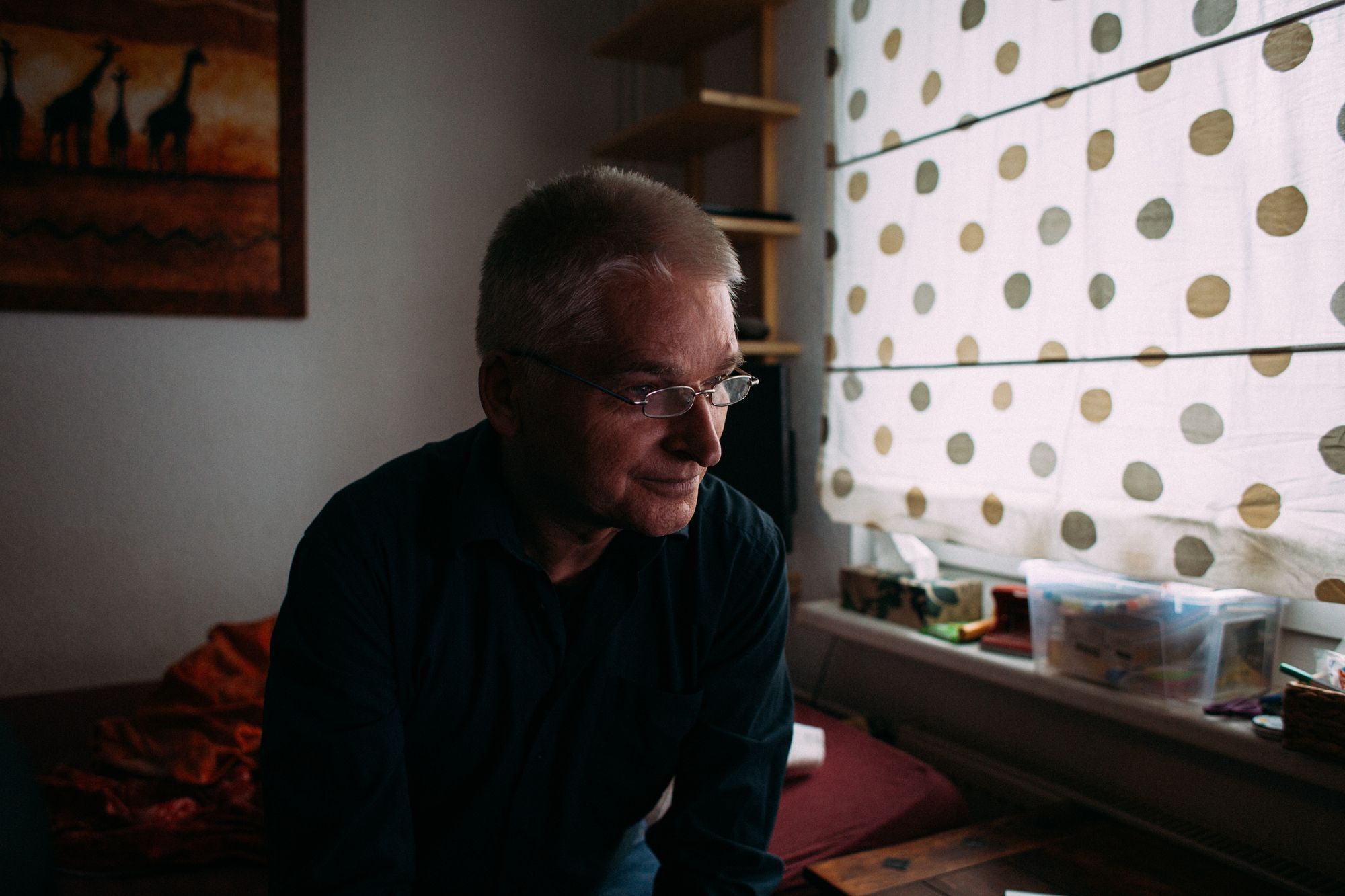 In einem Schlafzimmer sitzt Rainer auf dem Bett, der durchscheinende Vorhang ist zu. Er lehnt nach vorne und sieht nach unten rechts.