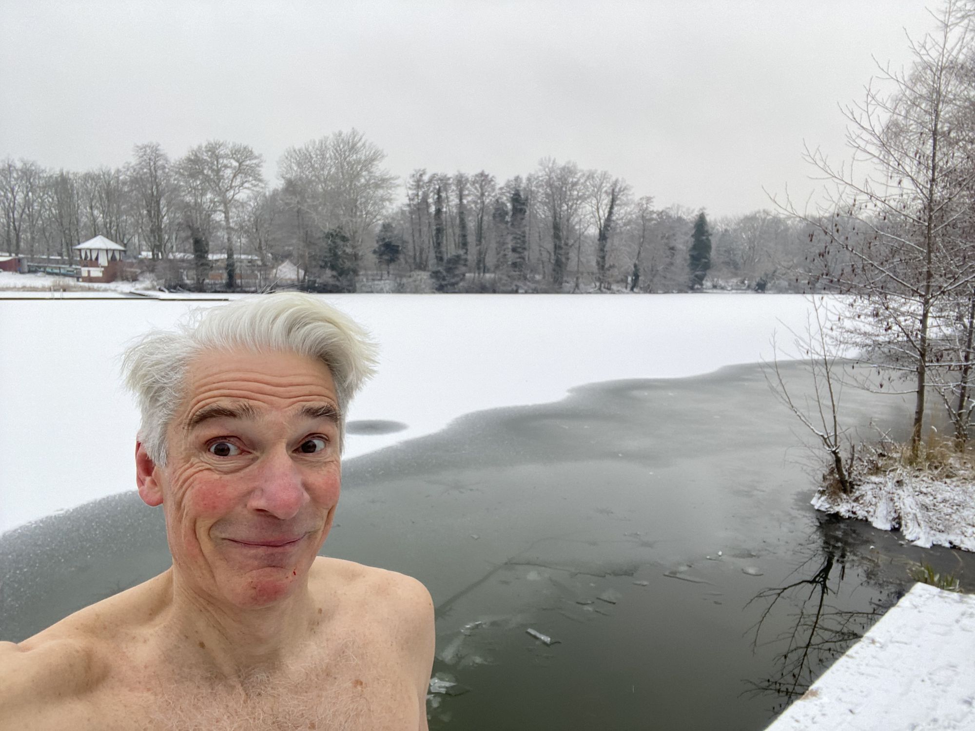 Man sieht einen älteren Mann, wie er mit naktemm Oberkörper freudestrahlend vor einem See steht, der in einer Winterlandschaft liegt.