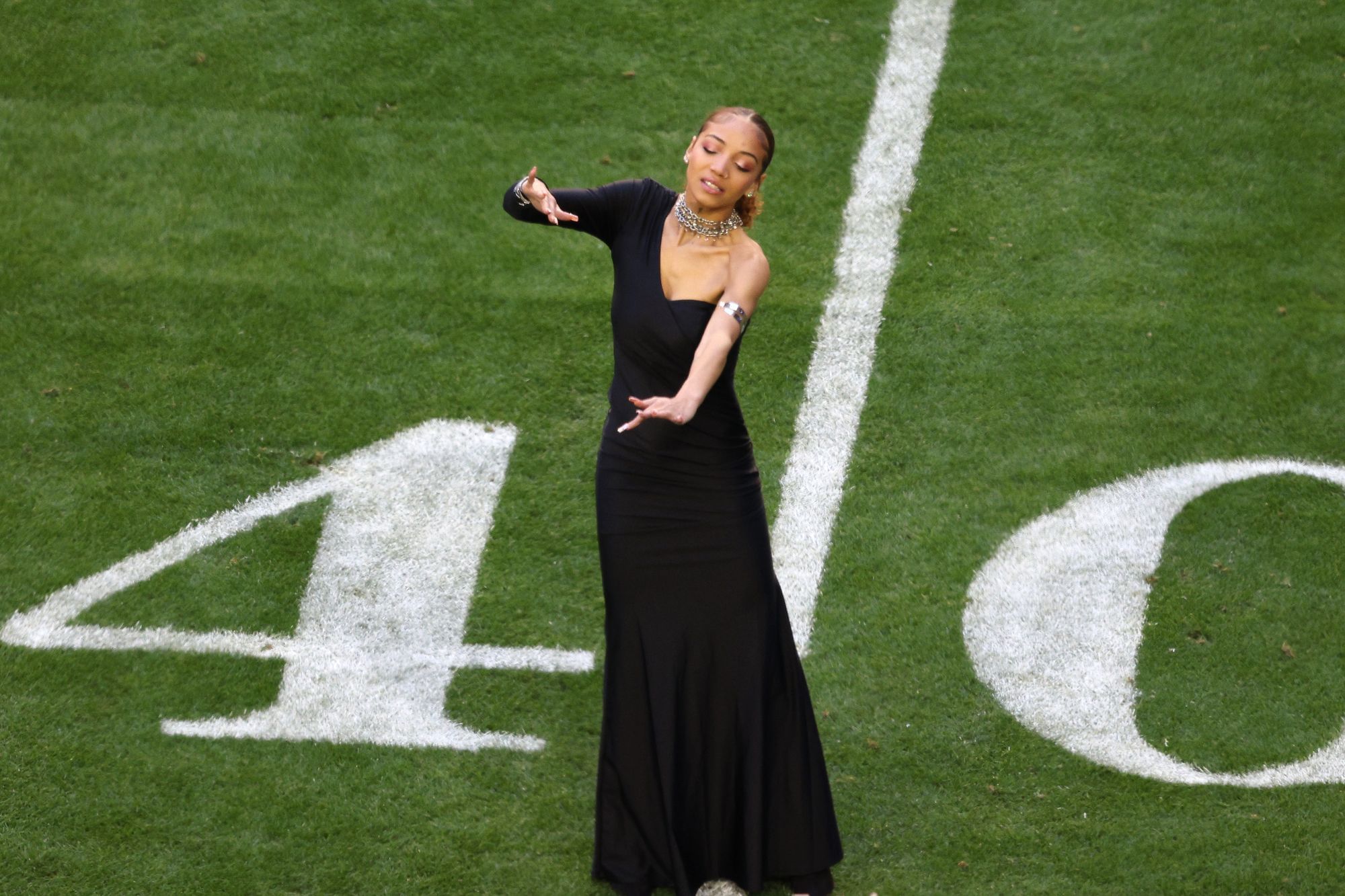 Man sieht eine schlanke Frau in einem asymmetrischen One-Shoulder-Dress in einem Stadion stehen, sie spricht in Gebärdensprache