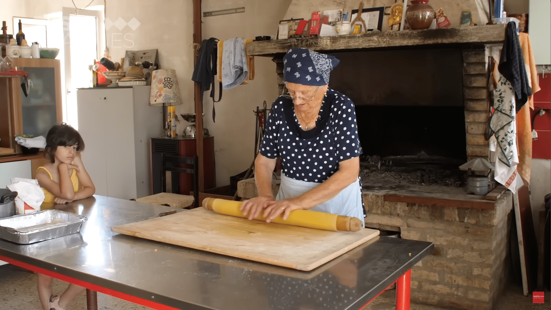Man sieht eine alte Dame, wie sie in einer Küche an einem großen Tisch einen Teig ausrollt. Es ist warm, denn die Frau trägt ein Tshirt, ein Kind, das neben dem Tisch steht und ihr zusieht, ein Trägertop. Im Hintergrund sieht man einen großen Steinofen.