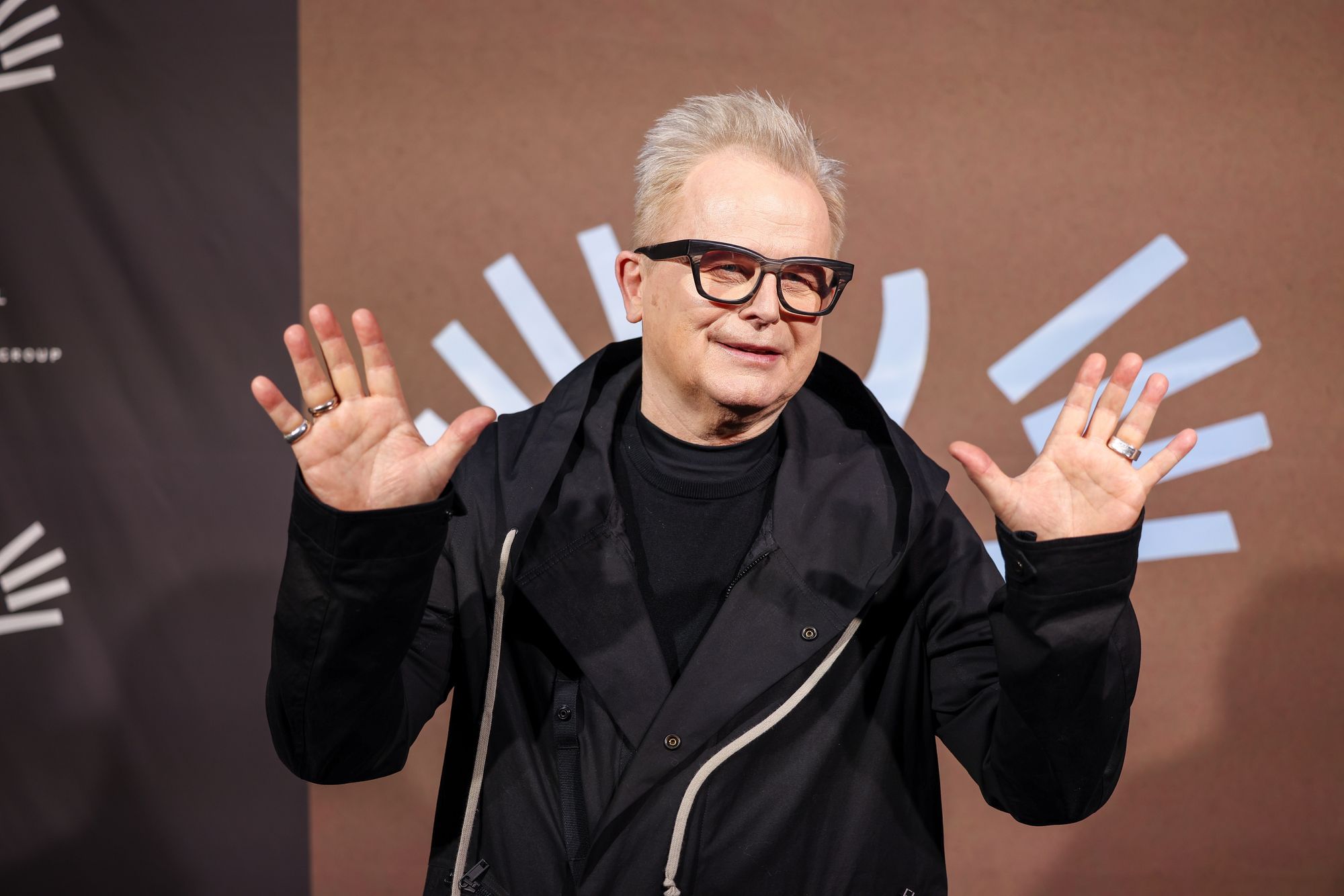 Man sieht den Sänger Herbert Grönemeyer, er trägt eine schwarze Statement-Brille, hebt beide Arme auf Schulterhöhe und öffnet die Handflächen als ein Winken in die Kamera.