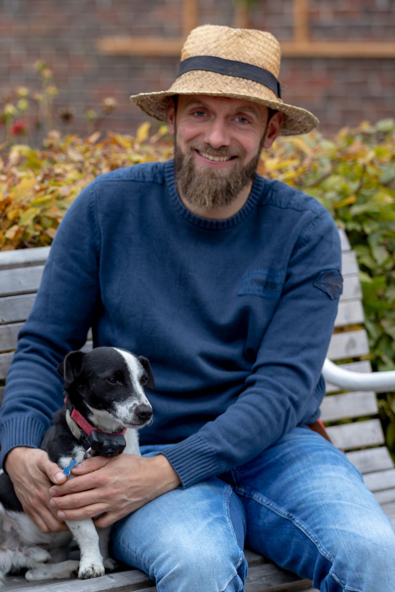Ein junger Mann mit Strohhut und Bart sitzt auf einer Holzbank und umarmt einen kleinen schwarz-weiß gefleckten Hund.
