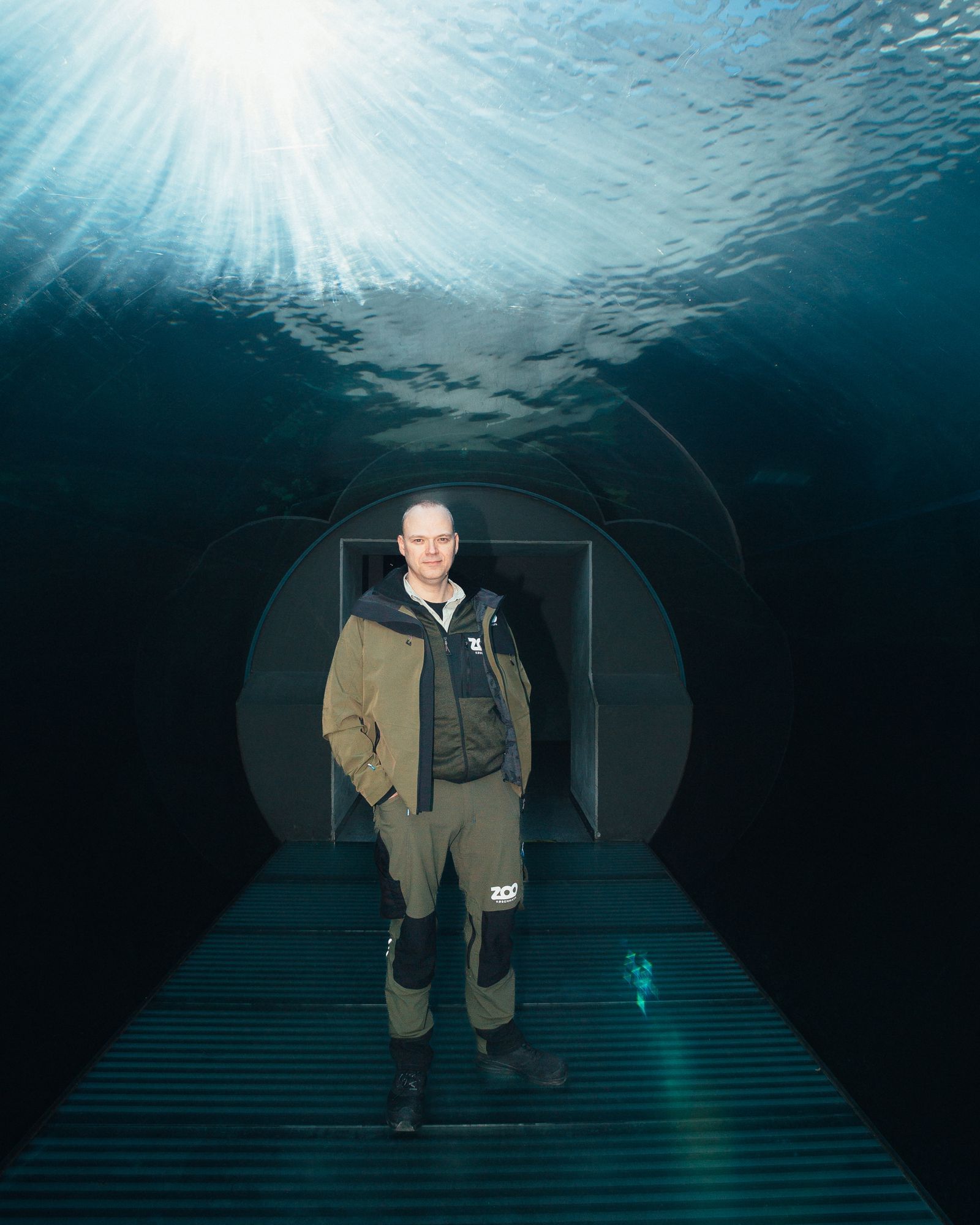 Ein Mann steht in einem gläsernen Tunnel unter Wasser. Von oben scheint die Sonne herein.