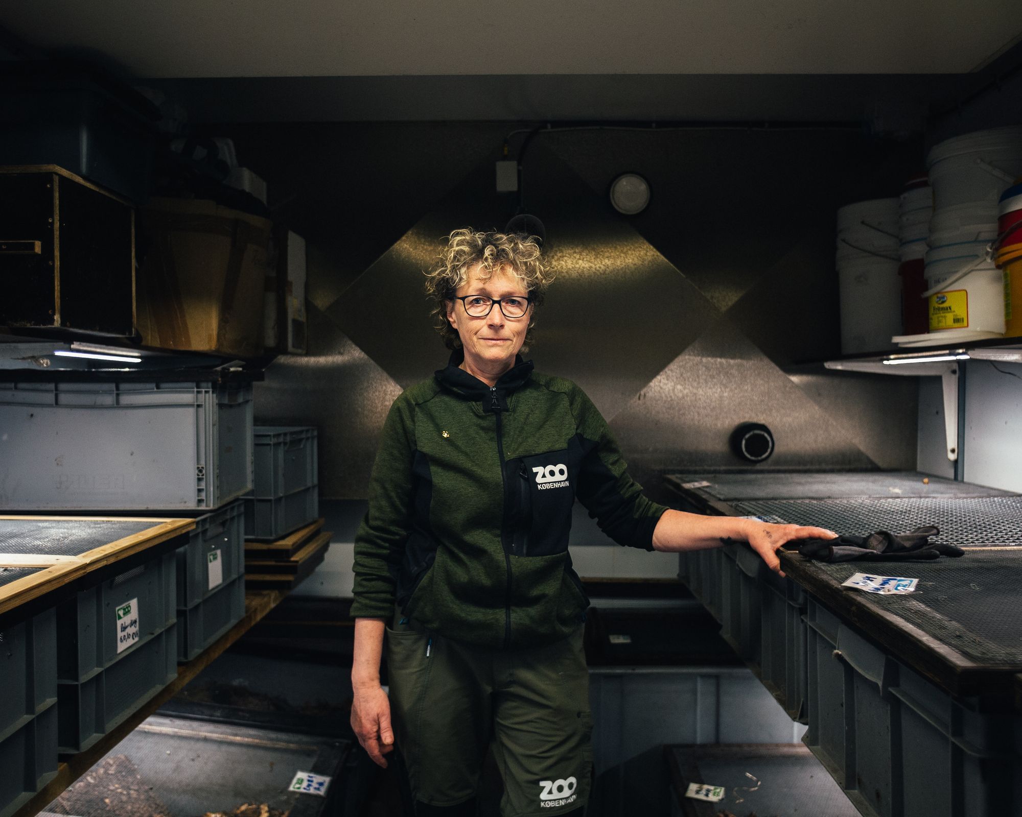Eine Frau mit kurzen blonden Locken steht in einem dunklen Raum mit grauen Plastikboxen