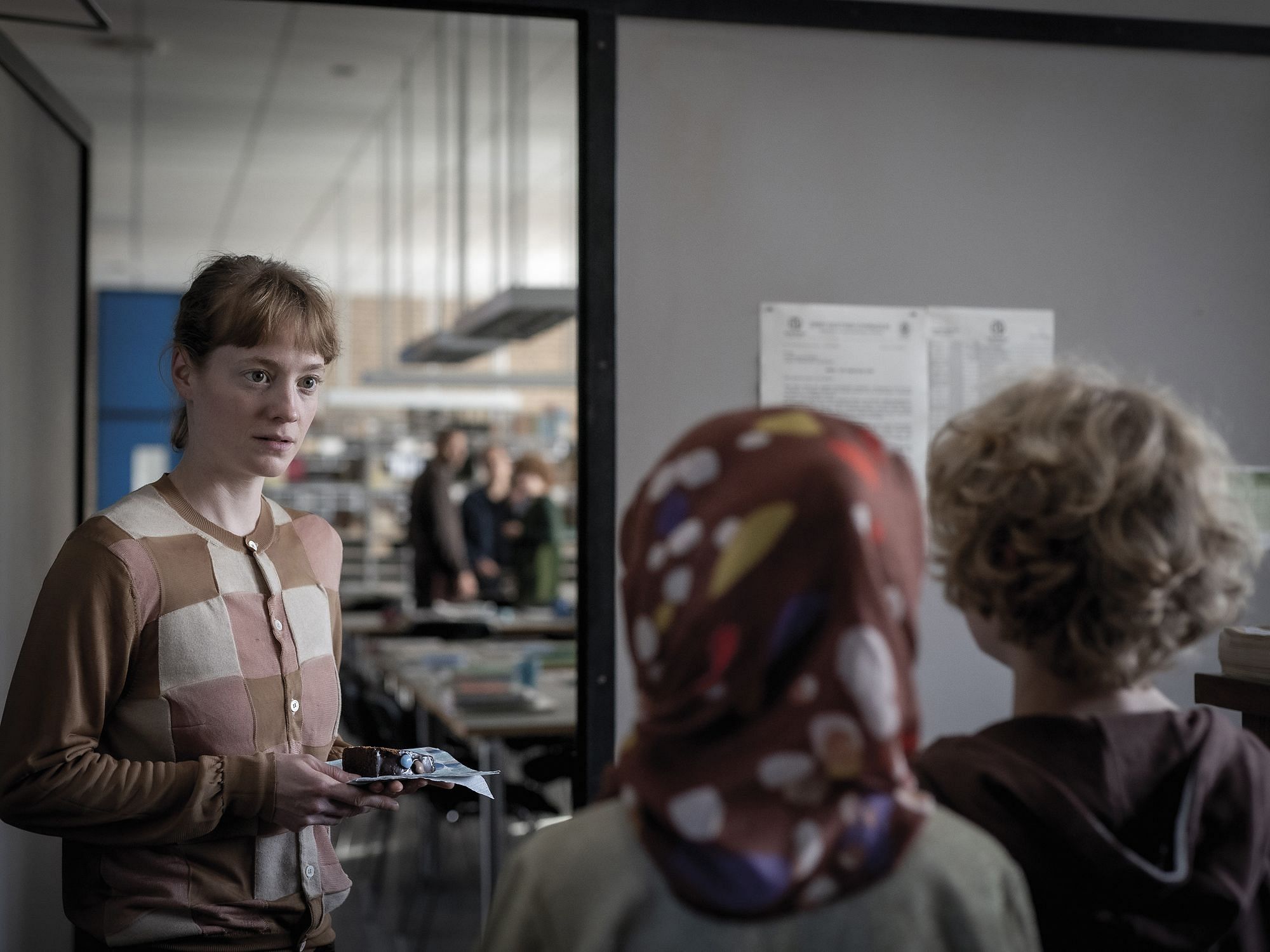 Die Hauptdarstellerin steht vor dem Lehrerzimmer und wird von zwei Schülerinnen angesprochen, die man nurn von hinten sieht.