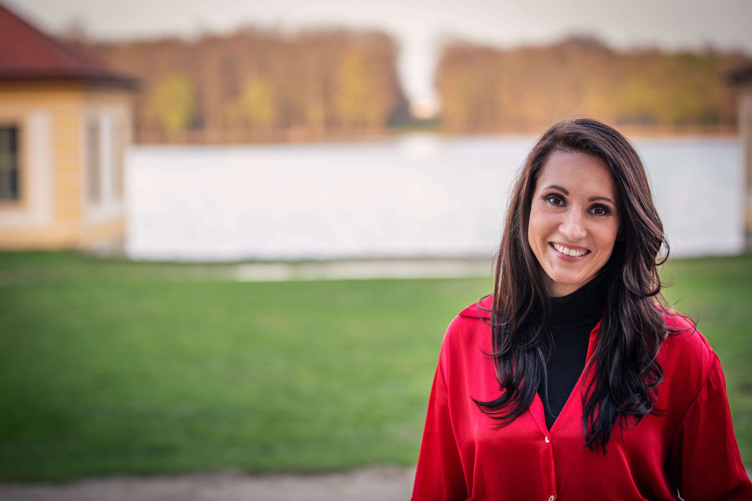Man sieht eine lächelnde Frau mit langen braunen Haaren, sie steht vor einem Fluss, im Hintergrund ist auch eine Wiese zu sehen, sie trägt einen schwarzen Rolli und eine rote Wollweste.