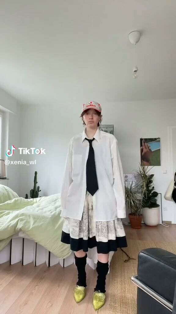 Stehende Person mit silberner Cap, weißem Oversize-Hemd, schwarzer Krawatte, schwarz-weißem Rock, schwarzen Socken und gelben, spitzen Schuhen