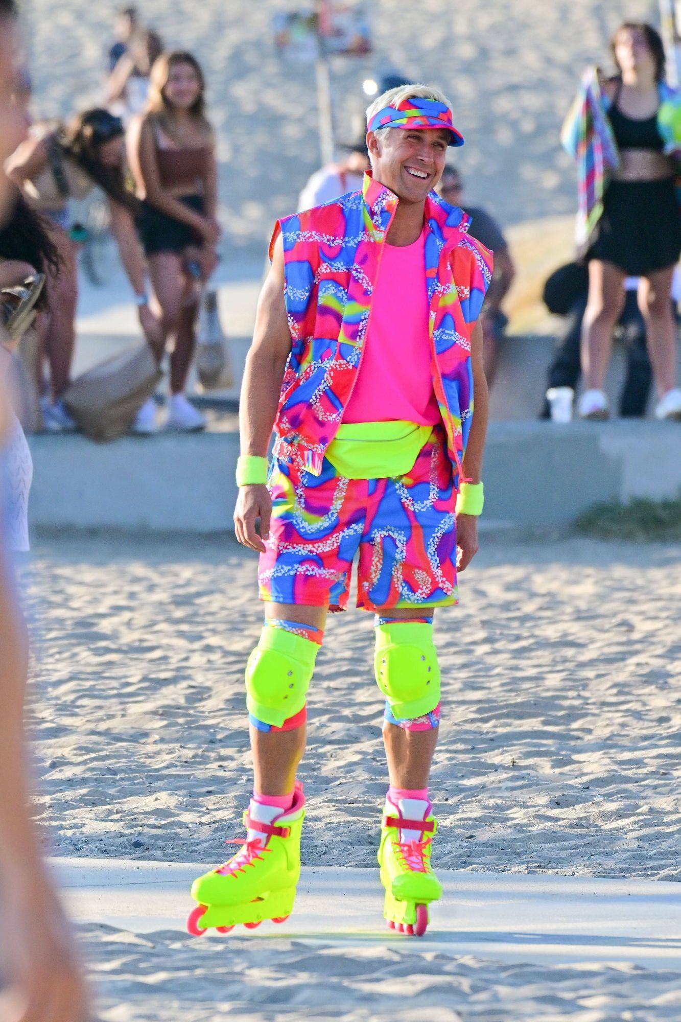 Man sieht den Schauspieler Ryan Gosling verkleidet in seiner Rolle als Ken in neon-gelben Rollerskates und neon-pinkfarbenem Outfit an einem Strand stehen, er lächelt breit.