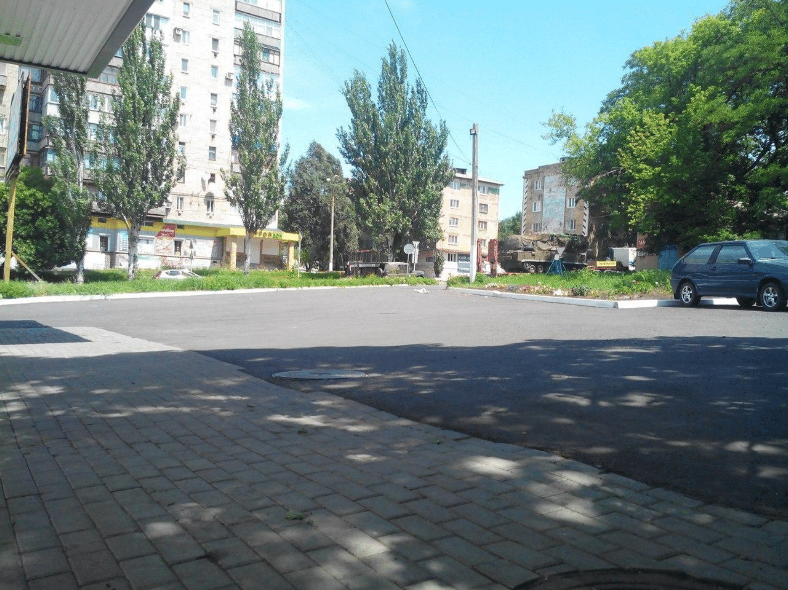Ein Platz in einer Wohnsiedlung, mit Schatten bedeckt. Rechts im Hintergrund: Ein grünes Militärfahrzeug mit dem Luftabwehrsystem.