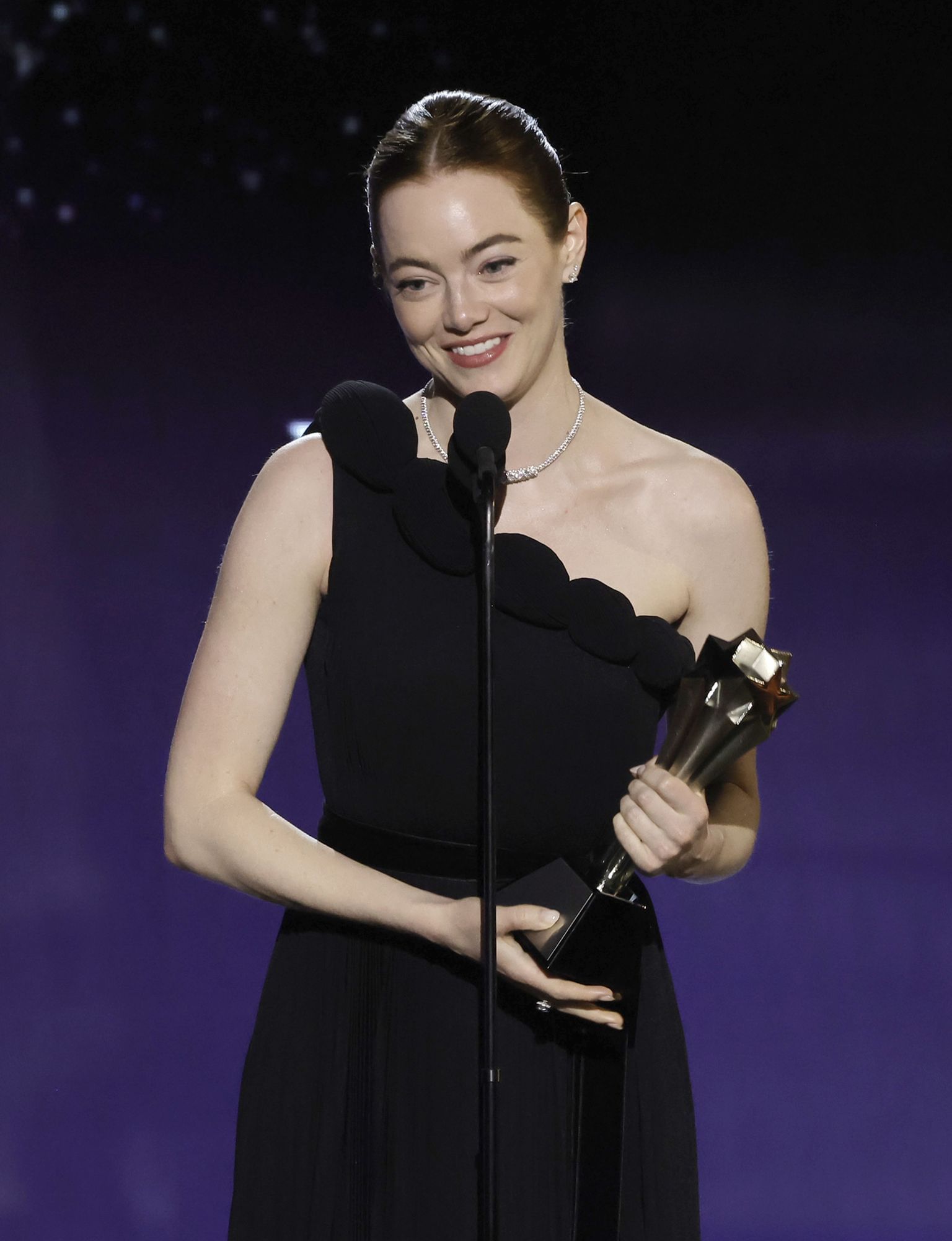 Man sieht die Schauspielerin Emma Stone in einem schulterfreien schwarzen Kleid bei einer Preisverleihung. Sie hält eine Trophäe in der Hand und spricht in ein Mikro, vermutlich ihre Dankesrede.