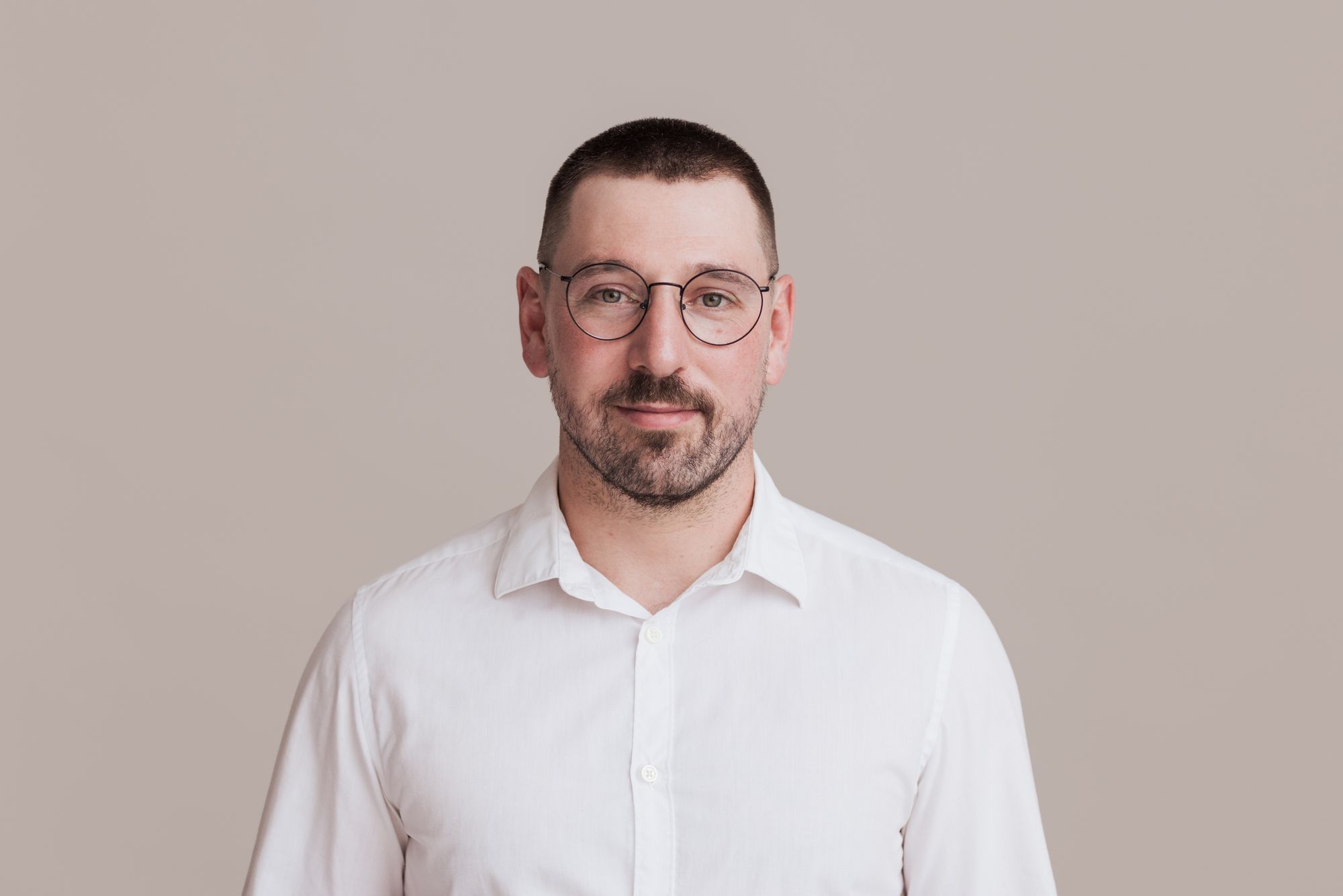 Auf dem Bild ist Anatol Bräunig, ein Psychologe, zu sehen. Er ist ein Mann mit kurzem dunkelblondem Haar und einem Vollbart. Er trägt eine runde Brille mit dünnem Metallrahmen und ein weißes Hemd. Der Hintergrund ist einfarbig und bietet einen kontrastarmen, neutralen Rahmen für das Porträt.