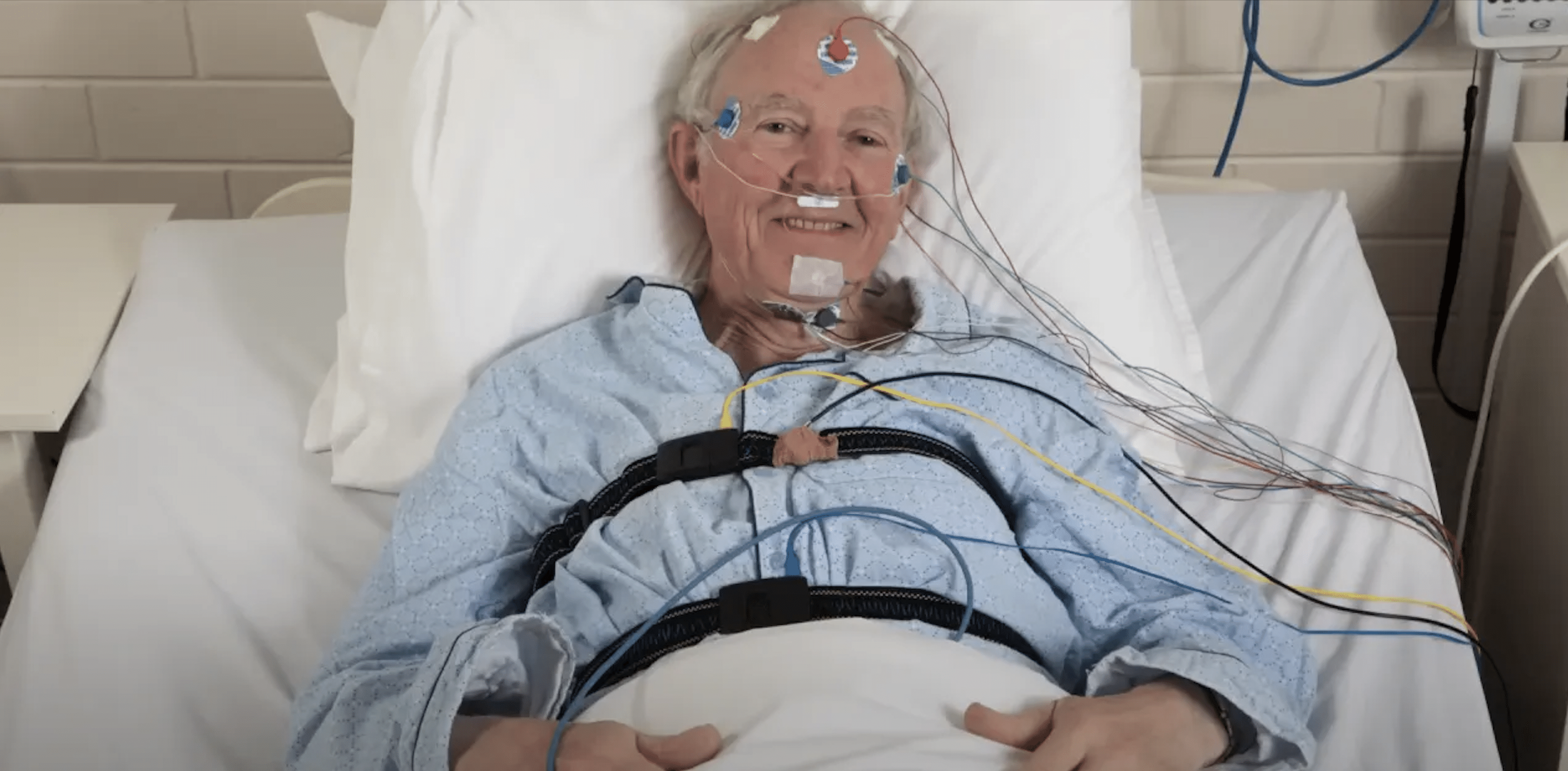 Ein Bild von einem Patienten im Schlaflabor, der an mehrere Schläuche angeschlossen ist. Mehrere elektrische Sonden sind in seinem Gesicht befestigt.