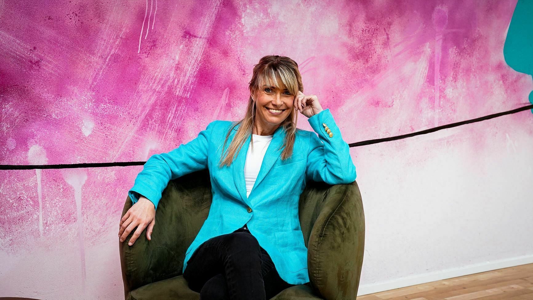 Auf dem Bild ist Anke Glasmeyer, eine Psychotherapeutin, zu sehen. Sie sitzt lächelnd und entspannt in einem olivgrünen, samtigen Sessel. Ihr Outfit ist lebhaft und professionell: ein türkisfarbener Blazer mit goldfarbenen Knöpfen am Ärmel, ein weißes Top und dunkle Hosen. Ihr blondes Haar fällt in weichen Schichten und sie hat einen freundlichen, offenen Gesichtsausdruck. Hinter ihr erstreckt sich eine lebendige, pinkfarbene Wand mit abstrakten weißen und dunkelrosa Farbspritzern. Ein schwarzes Kabel verläuft horizontal über die Wand und schafft einen markanten Kontrast zum Hintergrund.