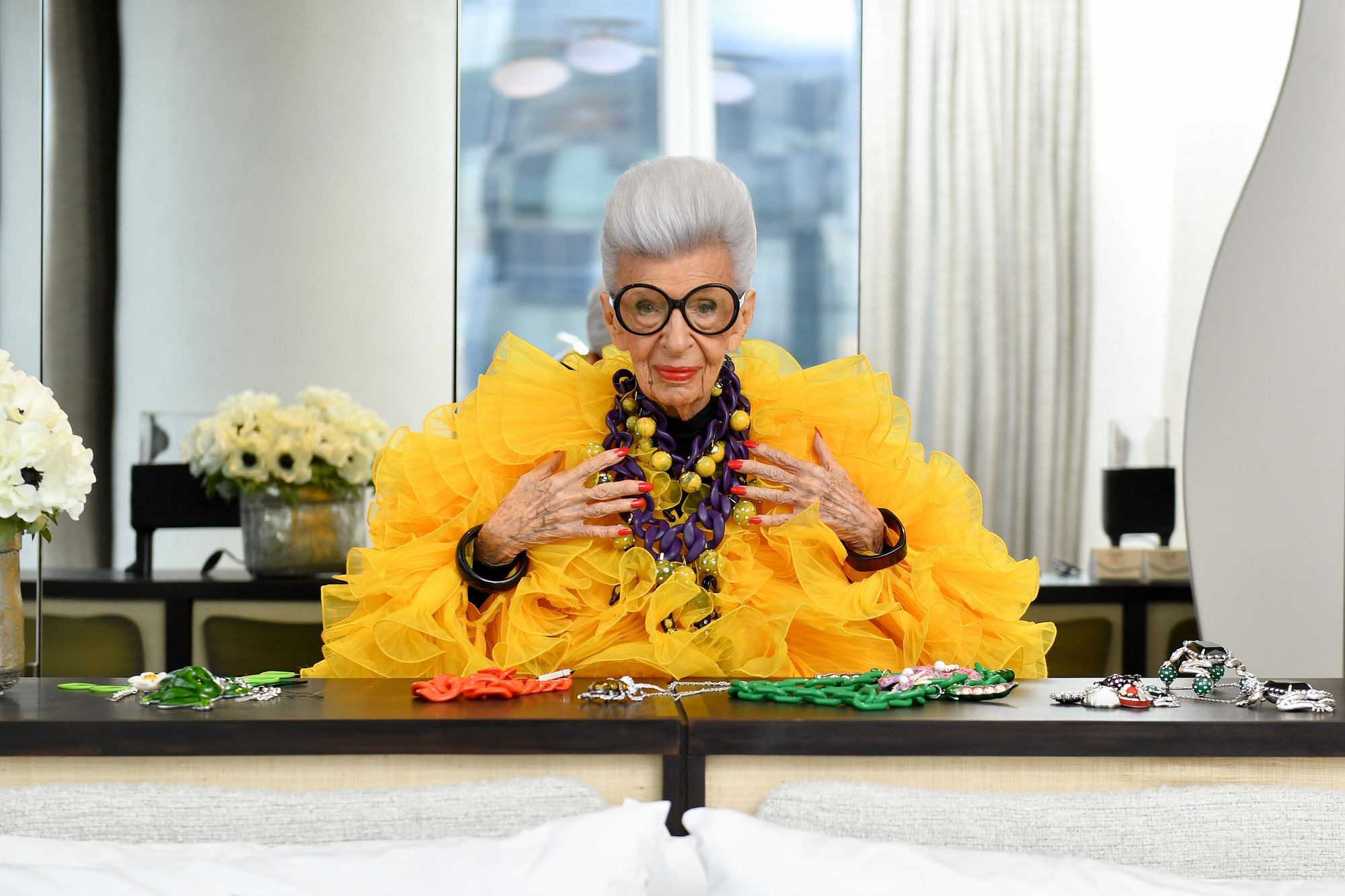 Iris Apfel sitzt an einem Tisch und trägt ein gelbes, fluffiges Oberteil.