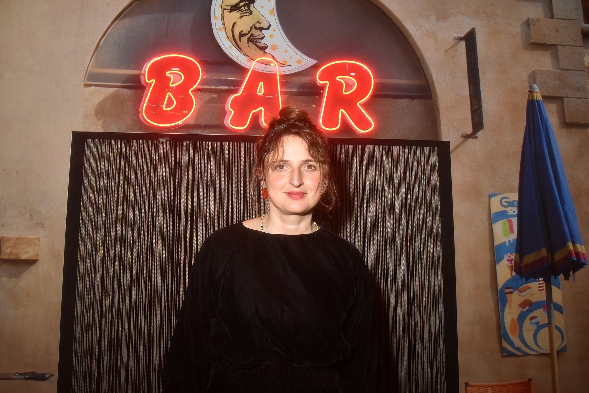 Alice Rohrwacher blickt in die Kamera, sie trägt ein schwarzes Oberteil. Hinter die leuchtet das Wort "Bar" in Neonschrift von einer Wand.