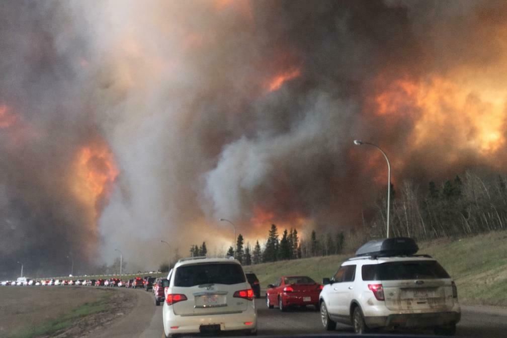Das Bild zeigt eine dramatische Szene, in der zahlreiche Autos auf einer Autobahn im Stau stehen, während sich rechts von ihnen ein großer Waldbrand ausbreitet. Dichter Rauch und Flammen sind deutlich sichtbar und färben den Himmel in bedrohliche Orange- und Grautöne. Der Verkehr scheint aufgrund der Nähe des Feuers vollkommen zum Erliegen gekommen zu sein, und die Situation wirkt äußerst gefährlich.