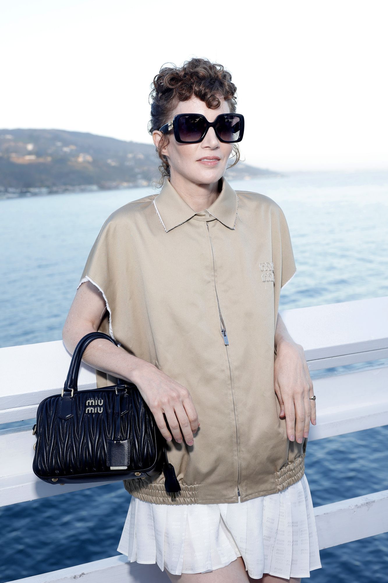 Miranda July steht an einer Brüstung, im Hintergrund sieht man das Meer. Sie trägt ein beiges Oberteil, einen weißen Rock und eine große, dunkle Sonnenbrille.