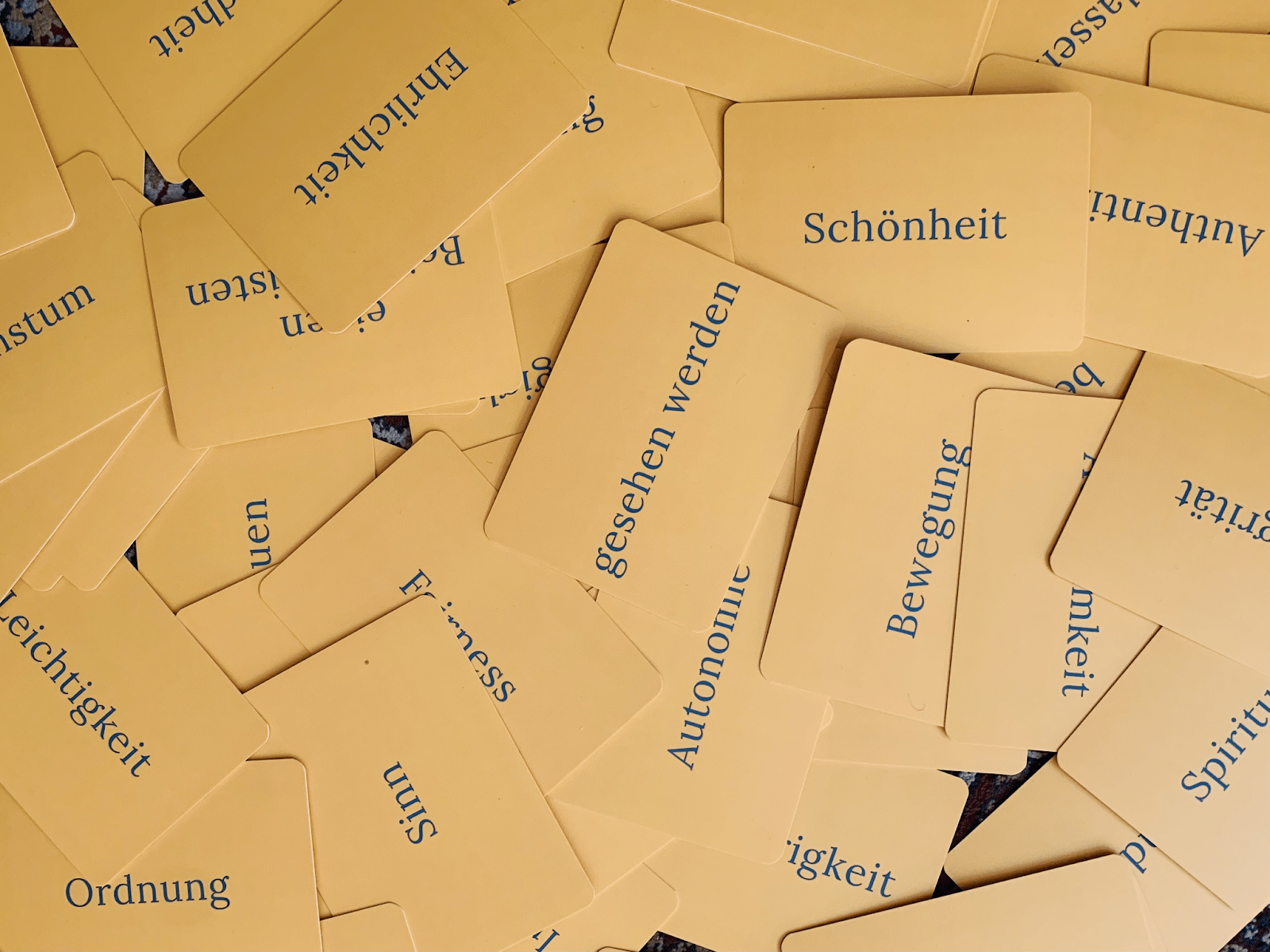 Auf dem Bild sind zahlreiche gelbe Karten zu sehen, die in scheinbar zufälliger Anordnung auf einer Oberfläche verstreut liegen. Jede Karte trägt ein anderes Wort, wie zum Beispiel Schönheit, Bewegung, Ordnung, Autonomie und Fairness. Diese Worte vermitteln verschiedene Konzepte und Werte, die möglicherweise zur Selbstreflexion oder als Impuls für Diskussionen und Aktivitäten verwendet werden könnten.