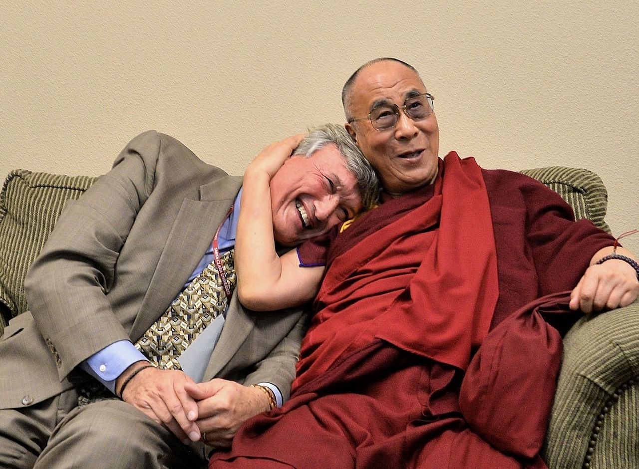 James Doty und der Dalai Lama sitzen auf einer Couch. James Doty lehnt seinen Kopf auf die Schulter des Dalai Lama.