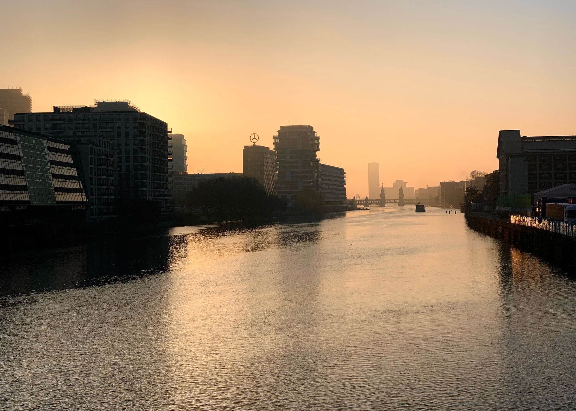 Auf dem Bild sehen wir einen Sonnenaufgang von der Schillingbrücke aus. Der Blick geht über die Spree in Berlin.
Die Sonne ist gerade über den Horizont gestiegen und taucht die Szene in ein warmes, goldenes Licht.
Rechts und links des Flusses stehen moderne Gebäude. Links sind vor allem Bürogebäude, rechts ist ein Gebäude mit einem großen Mercedes-Benz-Stern auf dem Dach erkennbar.
Im Hintergrund sieht man die Oberbaumbrücke mit ihren markanten Türmen.
Ein Boot fährt auf der Spree in Richtung der Brücke.