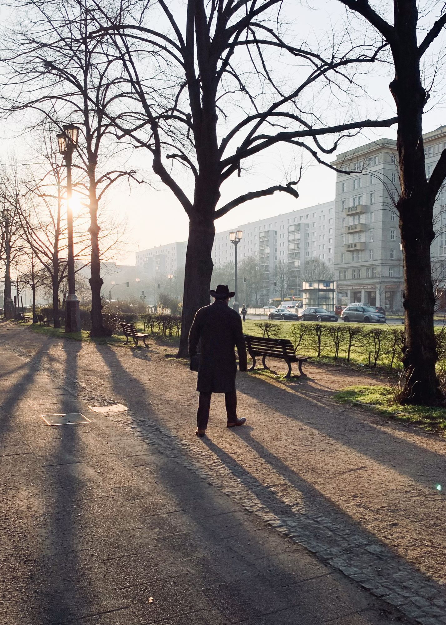 Das Bild zeigt einen Spazierweg in einer städtischen Grünanlage bei Sonnenaufgang. Eine Person steht mit dem Rücken zur Kamera, trägt einen dunklen Mantel und einen Hut. Die Sonne wirft lange Schatten, die sich über den Weg erstrecken. Links und rechts des Weges stehen kahle Bäume, einige Parkbänke sind zu sehen. Im Hintergrund sind mehrstöckige Wohngebäude, vermutlich Plattenbauten, und einige parkende Autos zu erkennen. Die Szenerie wirkt ruhig und die warme Morgenstimmung wird durch das weiche Licht unterstrichen.